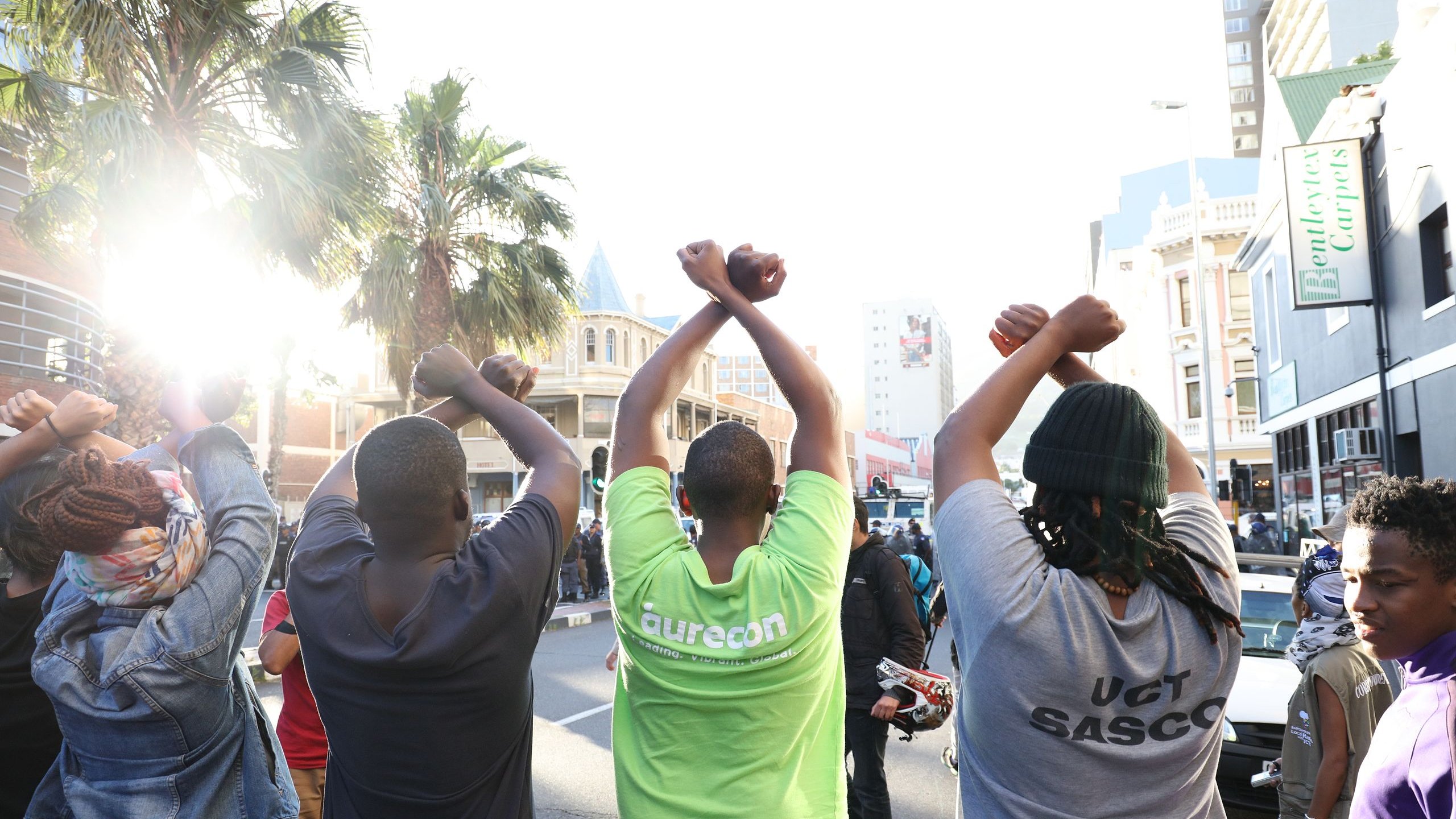 L'image montre un groupe de personnes vues de dos, levant les bras au-dessus de leurs têtes, formant un geste de solidarité ou de protestation. Ils sont sur une rue animée, entourés de bâtiments urbains. On peut apercevoir des palmiers à l'arrière-plan, ce qui ajoute une note de chaleur à l'environnement. La lumière du soleil éclaire la scène, créant une ambiance dynamique. Les personnes portent des t-shirts de différentes couleurs, dont l'un est vert vif. L'ensemble dégage une impression de union et d'engagement collectif.