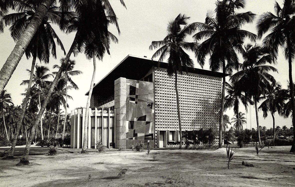 L'image montre un bâtiment moderne, avec une architecture distincte et géométrique. Le mur de la façade est ornée de motifs en relief, créant un jeu d'ombres et de lumière. Des colonnes blanches se dressent devant l'entrée principale, ajoutant une touche de classicisme à la structure. Autour du bâtiment, des palmiers élancés se balancent doucement, donnant une ambiance tropicale. Le sol est nu, avec une végétation assez faible, ce qui permet de mettre en valeur l'architecture. La scène semble baignée dans une lumière naturelle, évoquant une atmosphère calme et sereine.