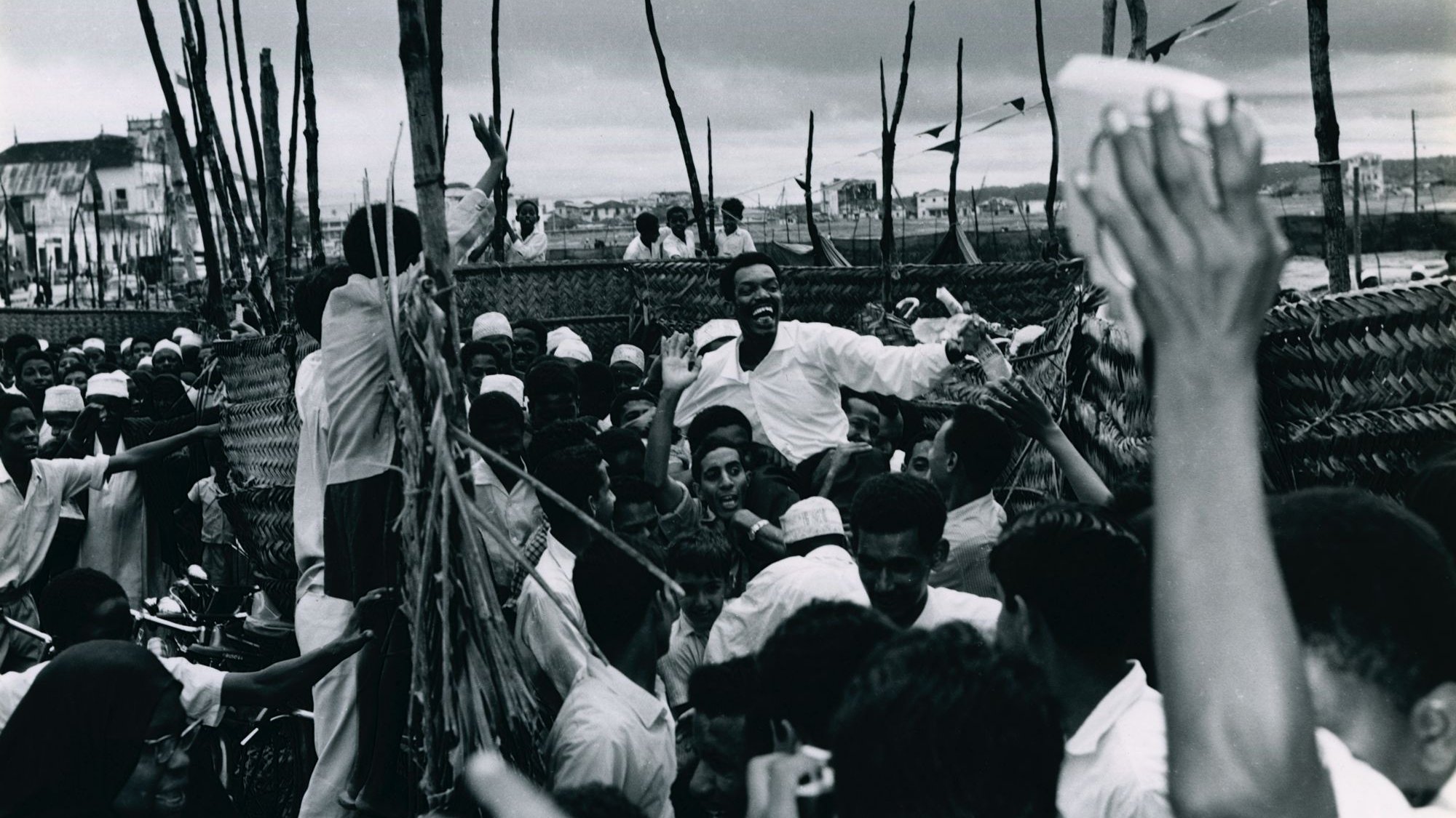 L'image montre une scène animée et dynamique, probablement lors d'un rassemblement ou d'une célébration. De nombreuses personnes se regroupent, leurs visages expriment la joie et l'excitation. On peut voir des hommes et des femmes, certains portant des vêtements traditionnels. Au centre, un homme semble chercher à atteindre un groupe, suscitant des réactions enthousiastes de la foule. Les bras de plusieurs personnes s'élèvent, clairement engagés dans l'événement. L'arrière-plan apparaît flou, avec des éléments de construction ou des arbres qui ajoutent à l'atmosphère vivante de la scène. L'ambiance générale évoque une forte connexion et une communauté vibrante.