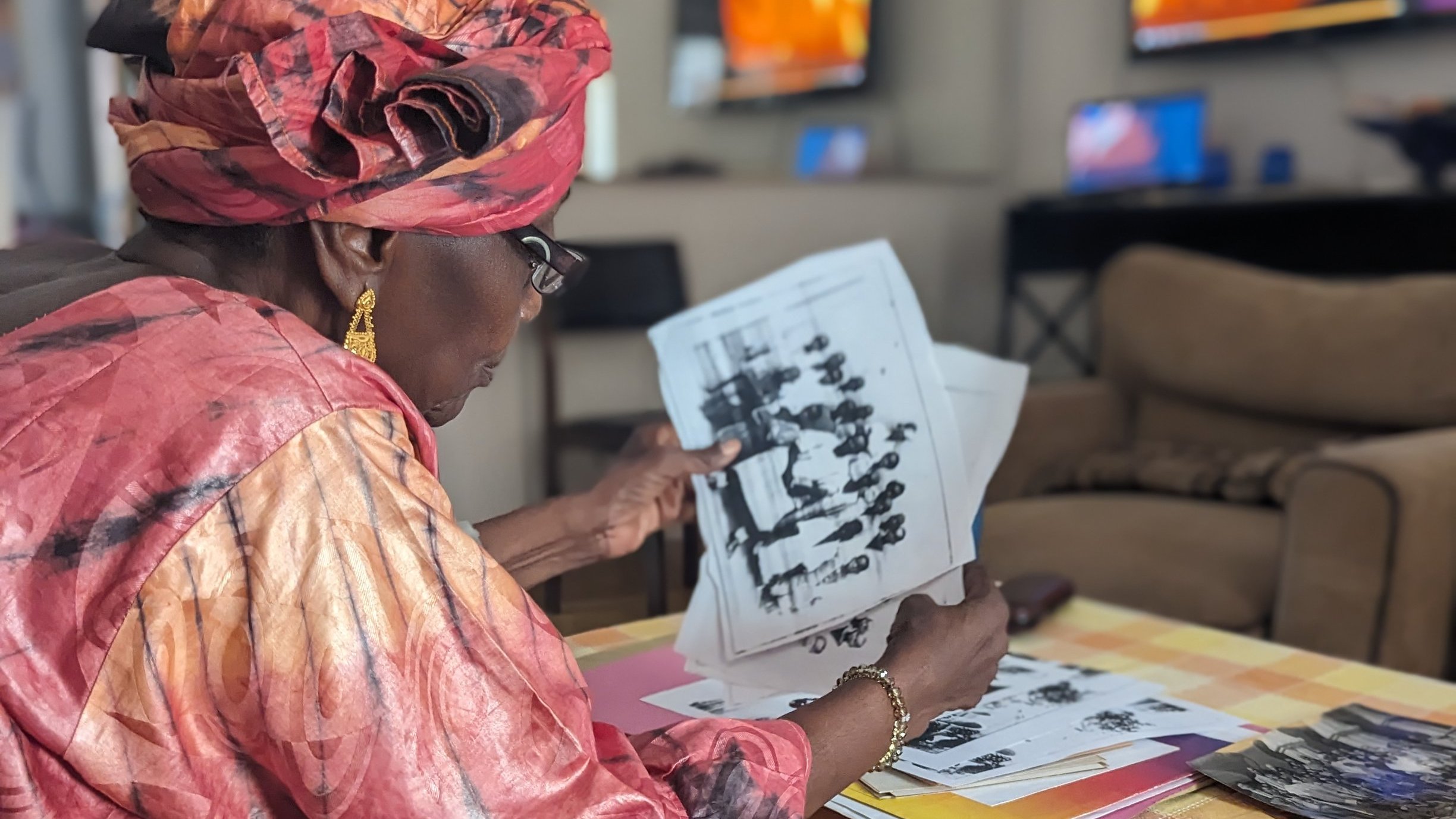 Dans cette image, une femme d'âge mûr, vêtue d'une magnifique robe rose et d'un foulard assorti, est assise sur un canapé dans un salon lumineux. Elle feuillette attentivement des photos en noir et blanc, toutes étalées devant elle sur une table basse recouverte d'une nappe à carreaux jaune et blanc. Le décor du salon est moderne, avec des télévisions visibles en arrière-plan, reflétant une ambiance chaleureuse et accueillante. La concentration de la femme témoigne de l'importance des souvenirs évoqués par ces images.