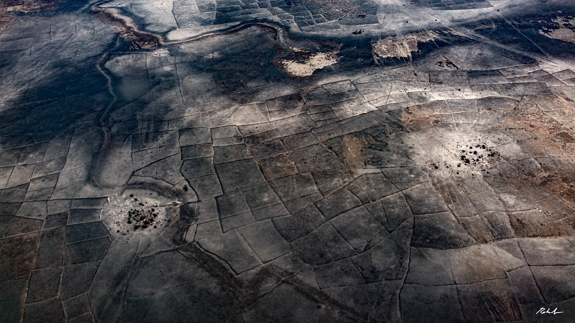 L'image montre un paysage vaste et aride vu d'en haut, avec des motifs géométriques formés par des parcelles de terre. Les teintes dominantes sont des nuances de gris et de marron, évoquant un terrain desséché, peut-être dans une région désertique. On y perçoit des zones plus sombres, qui pourraient représenter des végétations ou des matériaux différents. Les formes géométriques créent une texture visuelle intéressante, tandis que le sol semble craquelé par un manque d'eau. Cette scène dégage une sensation de vastitude et de tranquillité, tout en reflétant une nature rude et indomptée.