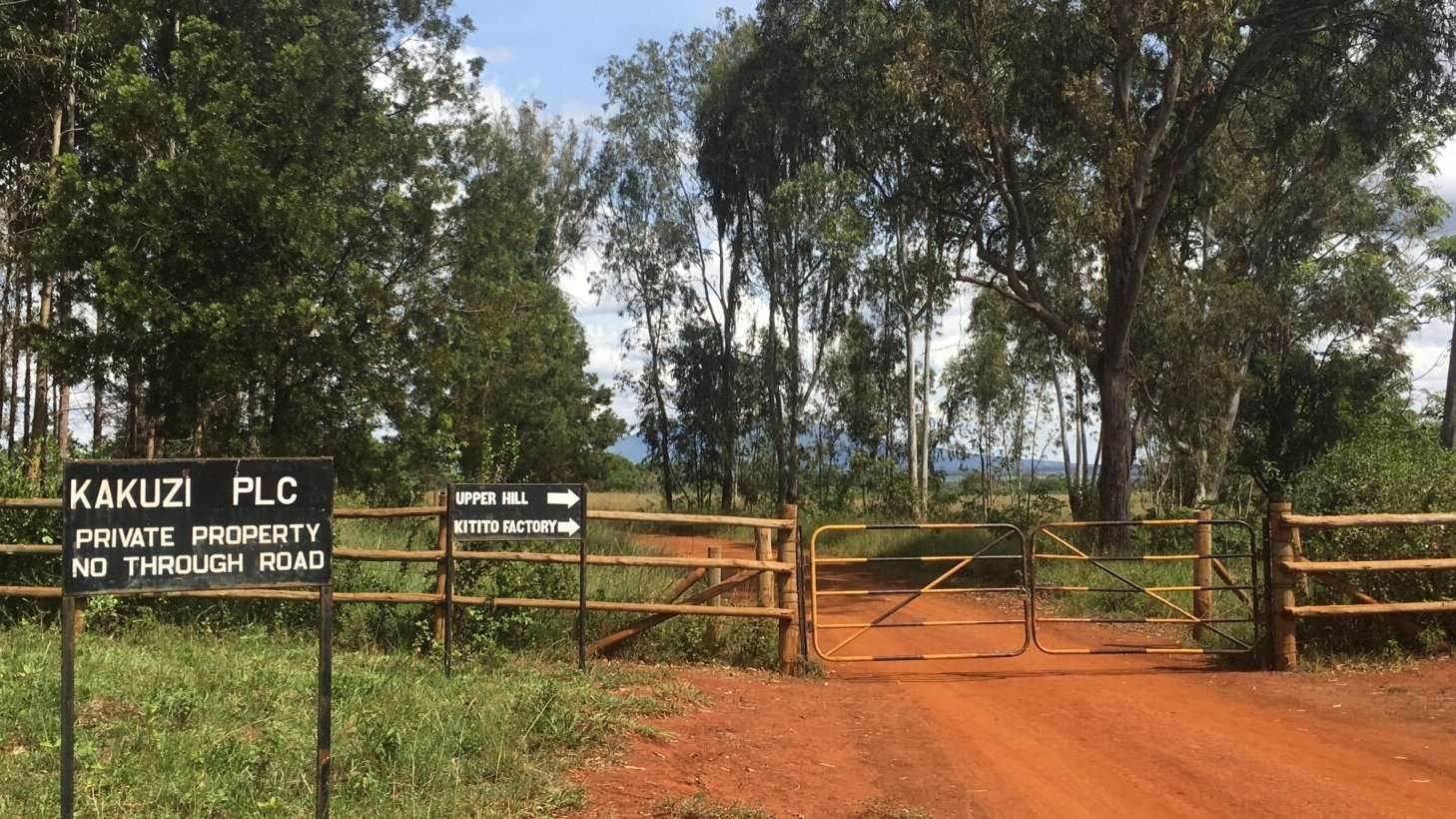 L'image montre une vue d'une rue d'accès menant à une propriété privée, entourée de végétation. À gauche, un panneau indique "KAKUZI PLC" et précise que c'est une propriété privée avec "NO THROUGH ROAD" (pas de passage autorisé). Le sol est recouvert de terre rouge, et une clôture en bois avec une porte fermée marque l'entrée. Derrière la clôture, on aperçoit des arbres feuillus qui encadrent le chemin. À droite, un panneau indique la direction vers "UPPER HILL" et une "KITTO FACTORY", ajoutant une touche d'orientation au paysage. Le ciel est clair avec quelques nuages épars.