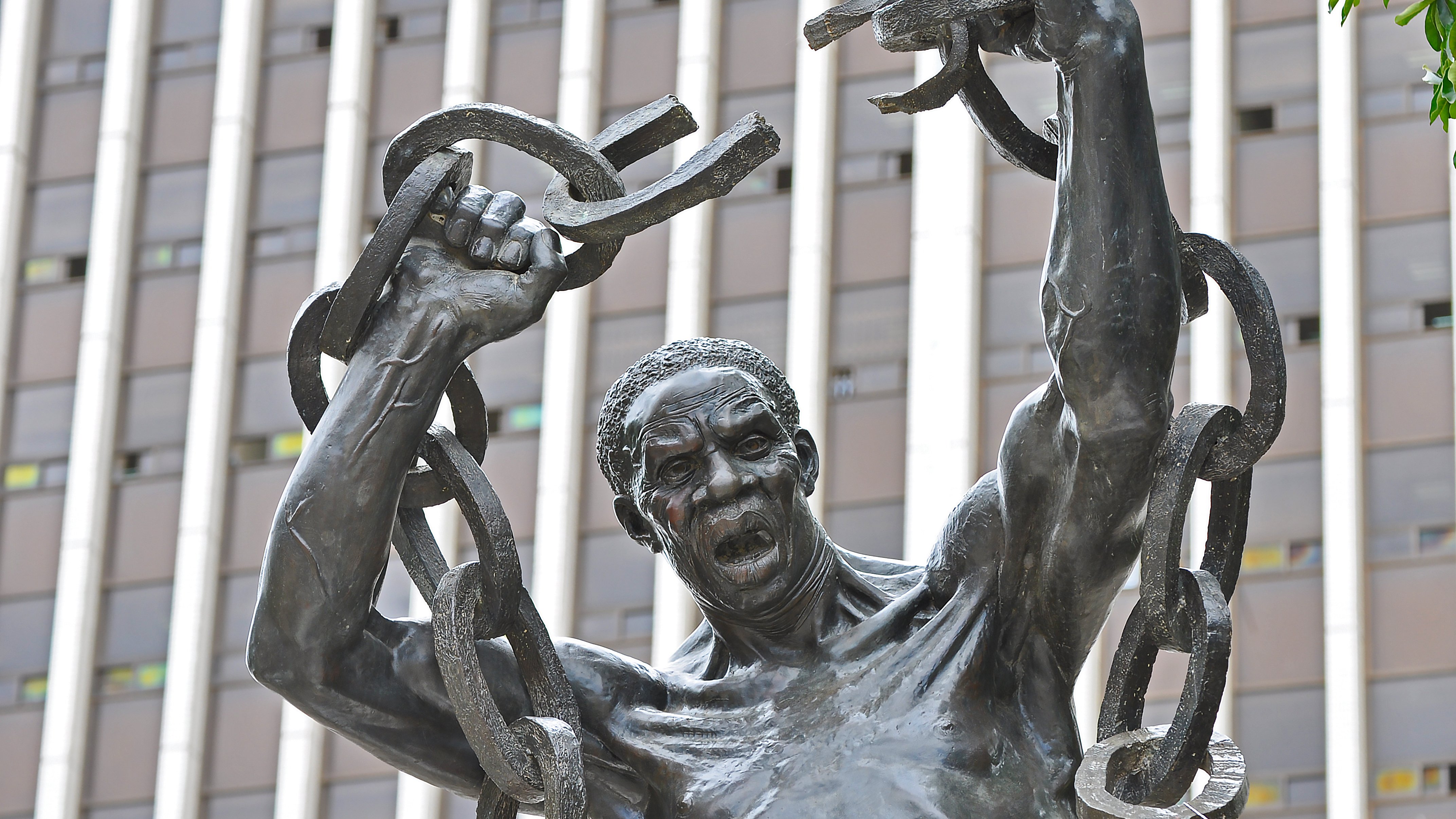 Cette image représente une statue en bronze d'un homme robuste. Il se tient debout, les bras levés au-dessus de sa tête, brandissant de lourdes chaînes brisées, symbolisant la libération et la force. Son visage exprime une intense émotion, avec une bouche ouverte comme s'il poussait un cri de victoire ou de défi. La sculpture est placée devant un bâtiment moderne, avec des lignes verticales qui soulignent contraste entre le passé et le présent. L'œuvre évoque des thèmes de résilience et de lutte pour la liberté.