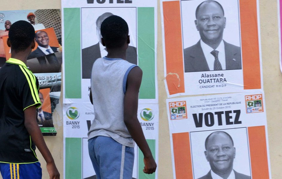 L'image montre un groupe de jeunes garçons marchant le long d'un mur recouvert d'affiches électorales. Les affiches présentent des portraits de candidats politiques, dont certaines affichent le nom d'Alassane Ouattara. Les affiches sont aux couleurs vives, avec des fonds colorés, principalement du rose et du vert, et des mots comme "VOTEZ". Les garçons, vêtus de t-shirts, semblent attentifs aux affiches. Le décor est urbain et évoque l'ambiance d'une campagne électorale animée.