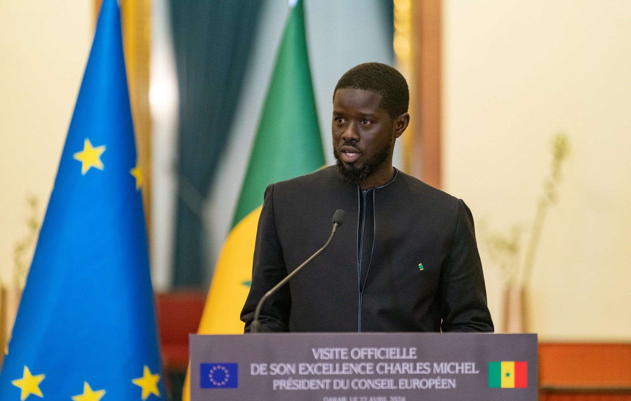 Sur cette image, un homme est en train de prendre la parole lors d'une conférence. Il est vêtu d'une tenue traditionnelle noire et se tient devant un podium portant une plaque avec des inscriptions officielles. En arrière-plan, on peut voir deux drapeaux : l'un affiche les étoiles de l'Union européenne et l'autre représente le Sénégal. L'homme semble sérieux et engagé, s'adressant à un public. La scène dégage une atmosphère formelle et diplomatique.