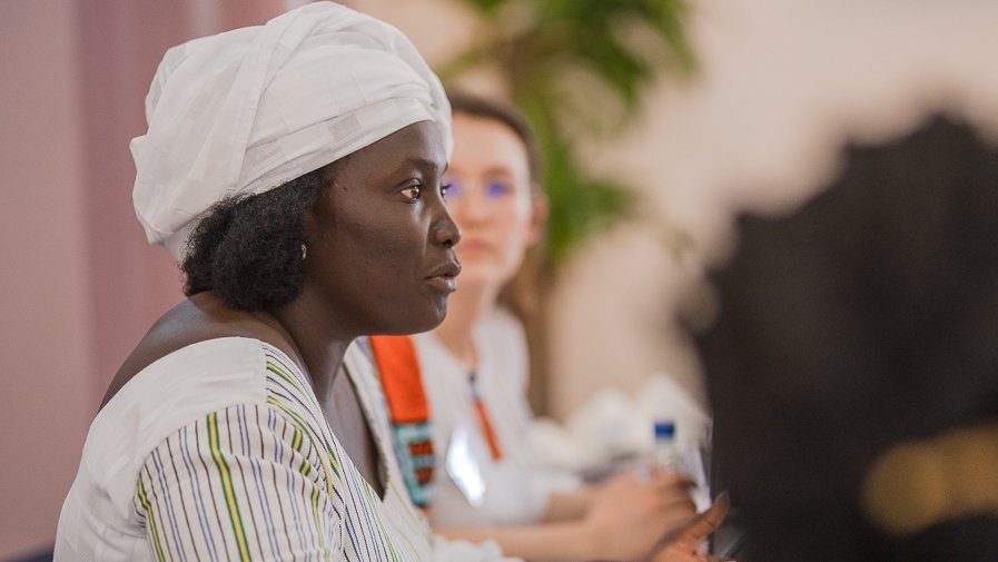 L'image montre une femme assise à une table lors d'une réunion ou d'une discussion. Elle porte une tenue traditionnelle, avec une chemise à rayures et un foulard blanc sur la tête. Son expression est sérieuse et concentrée, et elle semble engagée dans la conversation. À côté d'elle, une autre femme est présente, mais elle est floue en arrière-plan. On peut apercevoir des bouteilles d'eau sur la table, ainsi qu'une ambiance chaleureuse et collaborative. Des éléments de verdure, comme une plante, ajoutent une touche de couleur à la scène.