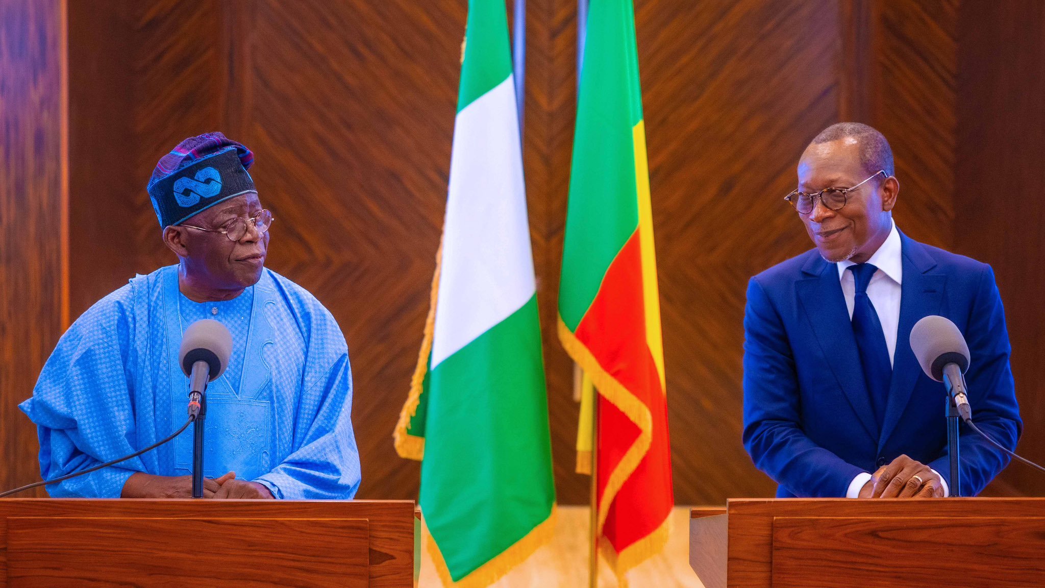 L'image montre deux hommes se tenant devant des pupitres en bois dans un cadre officiel. À leur arrière-plan, on peut voir les drapeaux du Nigéria (vert, blanc, vert) et du Bénin (rouge, jaune, vert). L'homme à gauche porte une tenue traditionnelle de couleur bleue avec un chapeau, tandis que l'homme à droite est habillé en costume sombre. Les deux hommes semblent engagés dans une conversation, exprimant une atmosphère de dialogue et de coopération. Le décor est élégant, soulignant le caractère officiel de la rencontre.