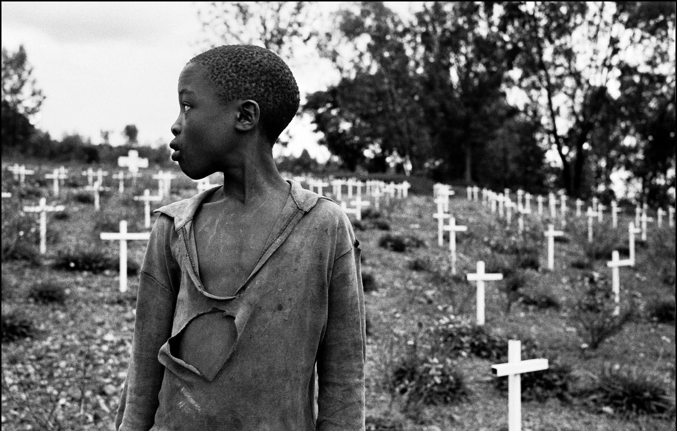 L'image présente un jeune garçon qui se tient dans un cimetière parsemé de croix blanches, symbolisant des tombes. Son regard est tourné vers la gauche, et on peut percevoir une expression de réflexion ou de tristesse sur son visage. Il porte un vêtement déchiré, soulignant une certaine vulnérabilité ou pauvreté. En arrière-plan, le paysage est entouré d'arbres, créant une atmosphère de mélancolie. Les croix blanches, disposées sur la pente, évoquent la mémoire et le souvenir de ceux qui ont perdu la vie.