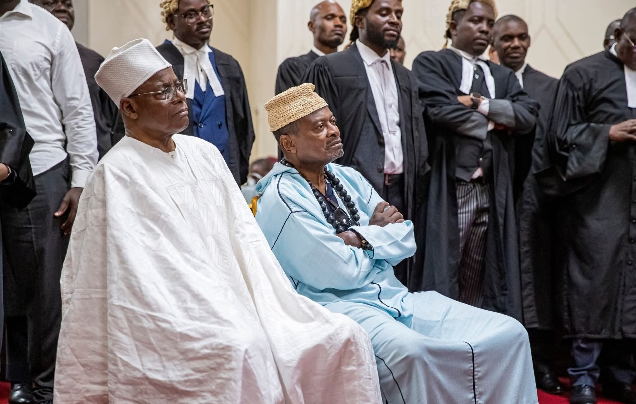 L'image montre un groupe de personnes rassemblées dans un espace formel, probablement un tribunal ou un événement solennel. Au premier plan, deux hommes sont assis. L'un porte une tenue traditionnelle blanche avec un chapeau, tandis que l'autre est habillé d'une tunique bleue et d'un collier de perles. Autour d'eux se trouvent des hommes en costume noir, certains portant des robes d'avocat. Tous semblent attentifs et sérieux, ce qui suggère que l'événement en cours est d'une grande importance. L'ambiance générale peut être perçue comme solennelle, avec une concentration visible sur les visages des participants.