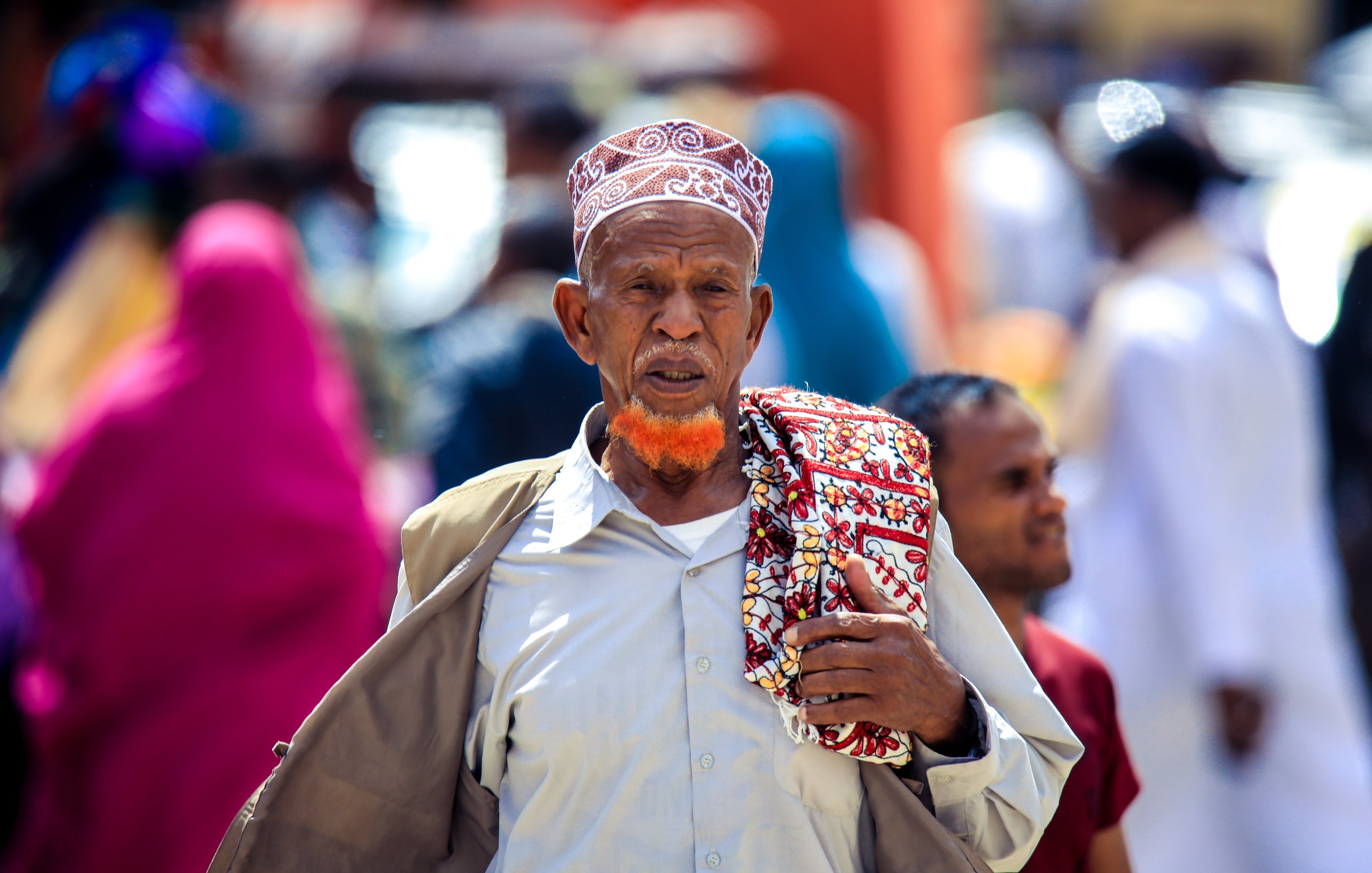 L'image montre un homme d'un certain âge marchant dans une rue animée. Il porte une tenue traditionnelle, avec une chemise claire et un grand châle coloré drapé sur son épaule. Sa tête est couverte d'un bonnet à motifs distinctifs, et il a une barbe teintée en orange, qui attire l'attention. En arrière-plan, on aperçoit des silhouettes floues de personnes vêtues de vêtements colorés, créant une ambiance vivante et dynamique. Les couleurs et la diversité des tenues témoignent d'un environnement culturel riche.