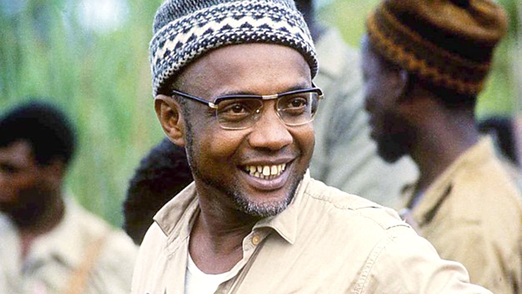 L'image montre un homme souriant, avec des lunettes au cadre fin et des cheveux rasés, portant une chemise beige. Il a une barbe grisonnante et son expression est chaleureuse et confiante. Il porte un bonnet tricoté, typique, dont les motifs évoquent un style traditionnel. En arrière-plan, plusieurs silhouettes floues d'autres hommes, habillés de manière similaire, suggèrent une ambiance de camaraderie et de travail en équipe. La scène véhicule une sensation d'optimisme et de détermination.