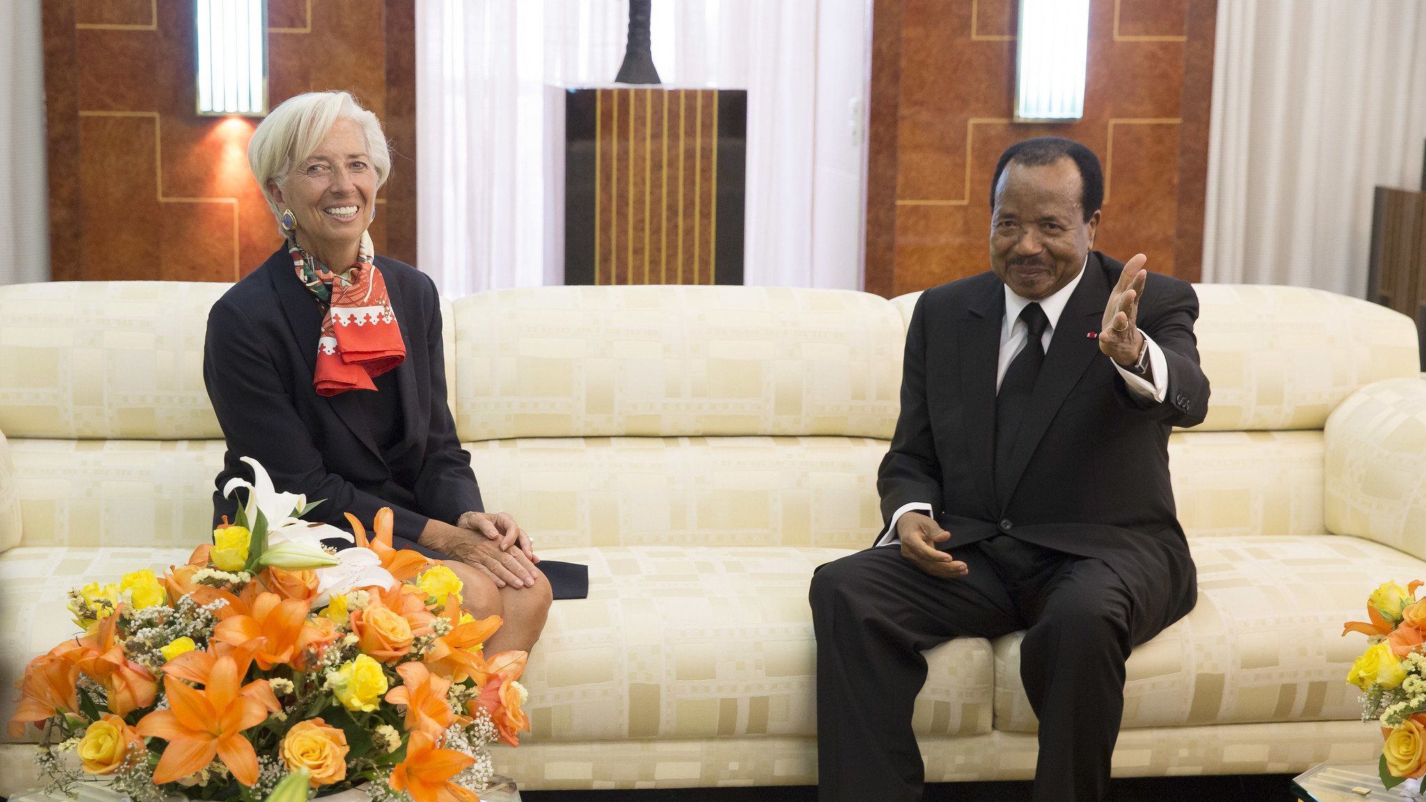 L'image montre deux personnes assises dans un salon élégant. À gauche, une femme aux cheveux courts et blonds, portant un blazer noir avec une écharpe rouge, sourit et semble détendue. À droite, un homme en costume noir et chemise blanche, assis sur un canapé crème, lève la main en signe de bienvenue ou pour parler. Ils sont entourés de bouquets de fleurs colorées, dominés par des teintes orange et jaune, qui ajoutent une touche de chaleur à l'environnement. Le décor est raffiné, avec des murs en bois et un éclairage doux, créant une ambiance conviviale.