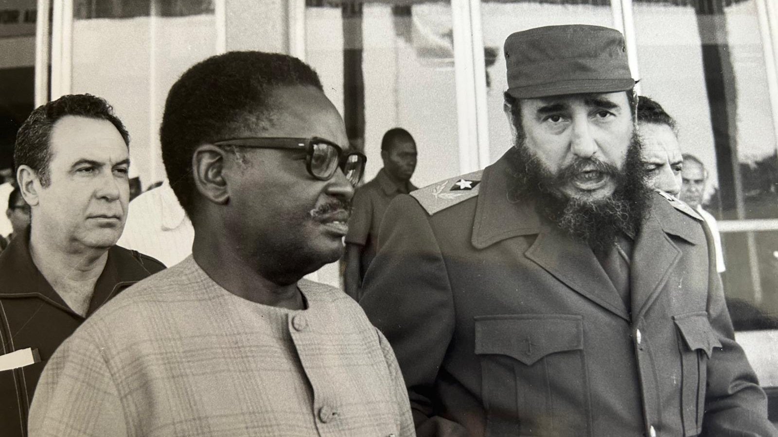 Cette image en noir et blanc montre trois hommes en discussion. Au premier plan, deux hommes sont particulièrement visibles. À droite, un homme avec une barbe fournie et un uniforme militaire, caractéristique de Fidel Castro, dialogue avec un homme portant des lunettes et un costume clair. Ce dernier semble attentif à la conversation. En arrière-plan, un troisième homme observe la scène. L'environnement est urbain, avec des bâtiments à vitre en arrière-plan. L'atmosphère paraît sérieuse et engagée, suggérant une rencontre diplomatique. Les expressions des visages laissent transparaître une certaine gravité.
