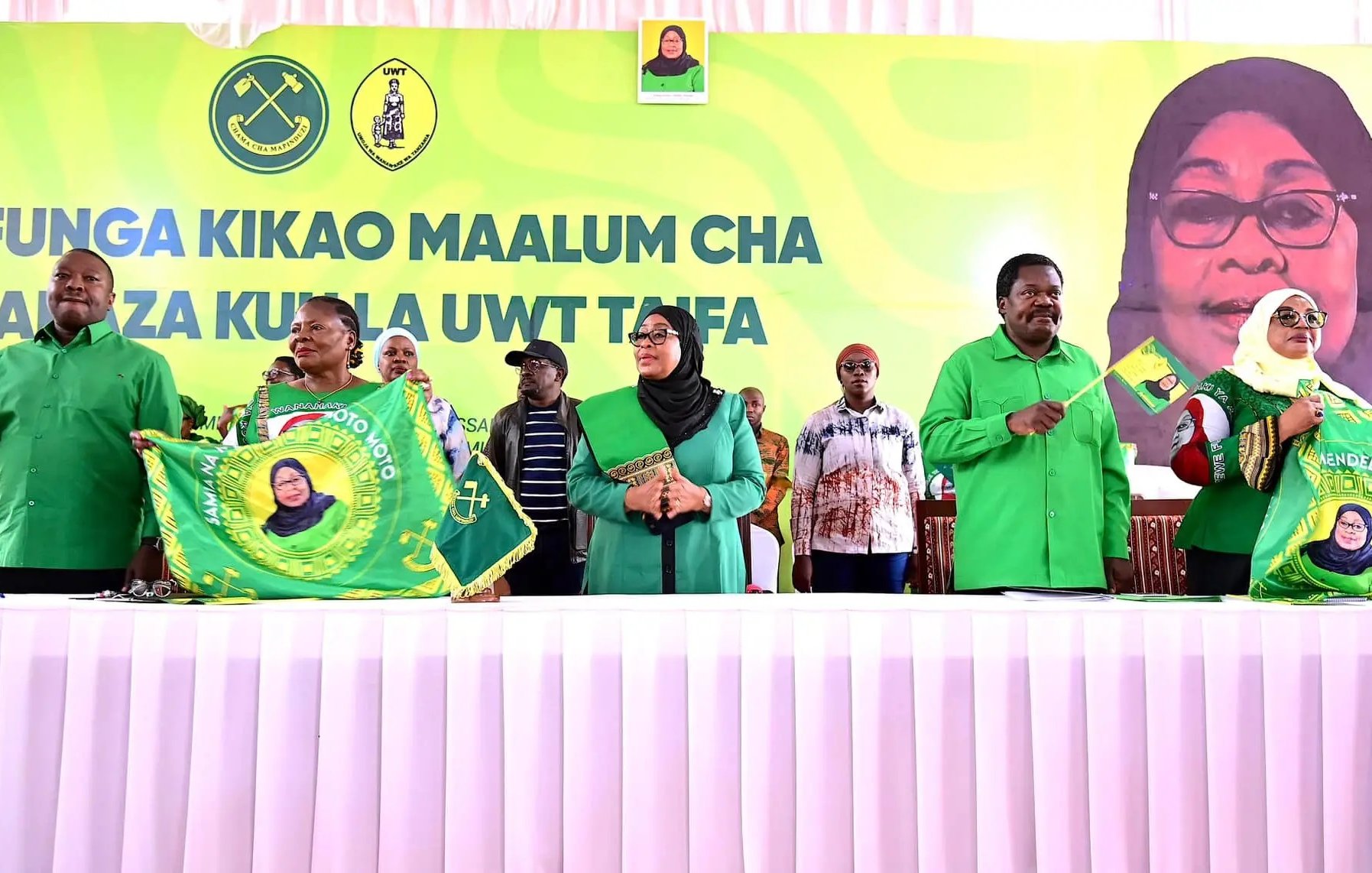 L'image montre un événement politique avec plusieurs personnes debout derrière une table, vêtues de chemises vertes. Au fond, on peut voir une grande bannière avec un portrait d'une femme souriante, probablement une dirigeante. Les participants tiennent des drapeaux ou des banderoles, souvent en vert et jaune, qui portent des symboles liés à leur organisation. L'atmosphère semble festive et engagée, avec des membres du public en arrière-plan qui écoutent attentivement. La réunion semble évoquer des thèmes de solidarité et de mobilisation.