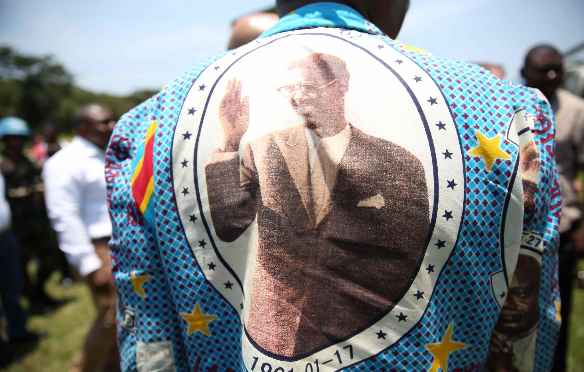 Cette image montre une personne portant un grand vêtement aux motifs colorés et vifs. Le dos du vêtement est orné d'une grande impression d'une personne en costume, levant la main dans un geste de salut. Autour de l'image, on peut voir des étoiles et des motifs géométriques. La date "1961-01-17" est inscrite, indiquant probablement une signification importante liée à l'image. En arrière-plan, on aperçoit un groupe de personnes, suggérant un événement ou une réunion en extérieur sous un ciel bleu.