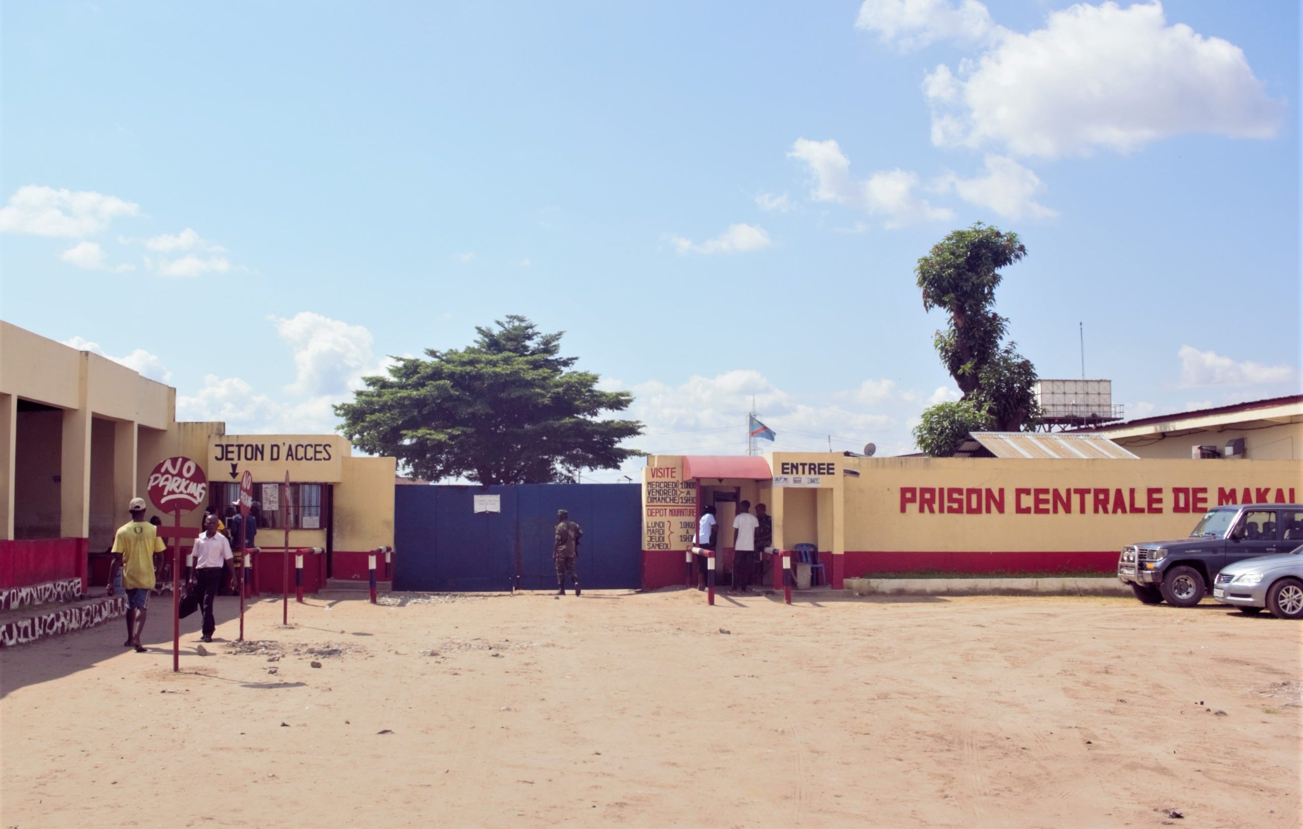 L'image montre l'entrée de la Prison Centrale de Makala. Devant le bâtiment, on voit une grande porte bleue. À droite, une cabine d'accès avec des petits guichets, probablement pour les visiteurs. Des personnes sont sur le point d'entrer ou de sortir. L'environnement est désertique, avec du sable au sol. Quelques voitures sont garées à droite, et un grand arbre se dresse à proximité. Le ciel est clair, avec quelques nuages. Des inscriptions sont visibles sur les murs, notamment le nom de la prison.