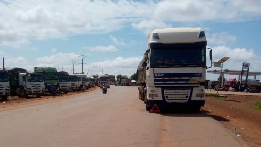 L'image montre une route large et droite, où une file de camions est stationnée sur le côté. Les camions, de différentes tailles et couleurs, sont alignés de manière ordonnée. Le ciel est clair avec quelques nuages blancs qui flottent. On peut apercevoir des poteaux électriques le long de la route, et au loin, il y a des bâtiments, suggérant un environnement urbain en arrière-plan. À proximité, un motocycliste passe, ajoutant un peu de dynamisme à la scène calme. L'ambiance générale dégage une impression de circulation routière et d'activités commerciales.