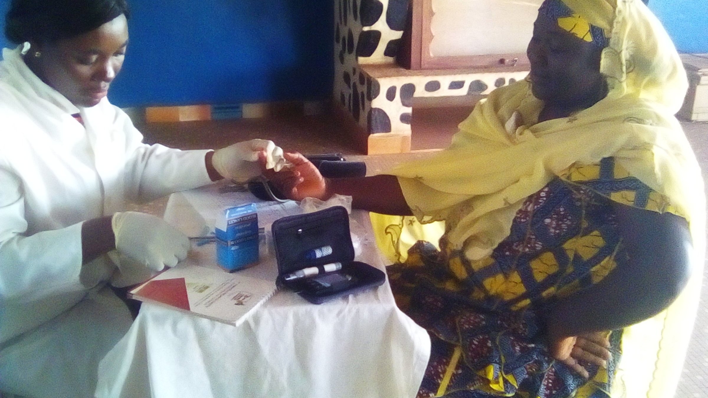L'image montre une scène dans une salle de consultation. À gauche, une professionnelle de la santé, vêtue d'un manteau blanc et de gants, effectue un test sur le bras d'une patiente. La femme qui reçoit le soin porte un turban jaune et une robe aux motifs colorés. Elle semble concentrée et un peu nerveuse. Sur la table devant elles, on peut voir des documents et des instruments médicaux, comme un petit appareil de mesure. Les murs de la pièce sont peints en bleu, et le sol est en béton. L'environnement est calme, et l'atmosphère est professionnelle, axée sur le soin et le soutien.