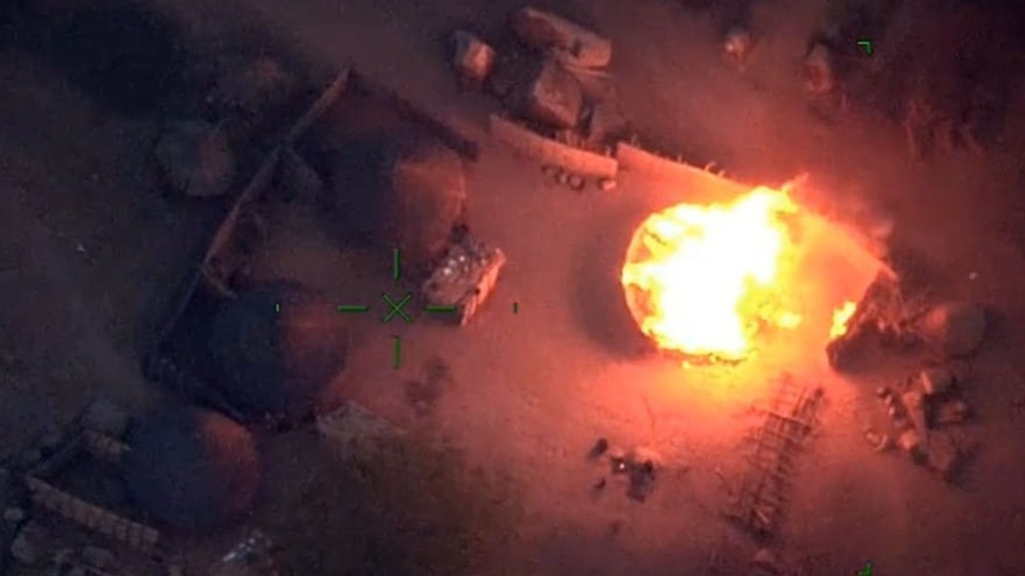 L'image représente une scène vue du ciel, où l'on aperçoit un environnement rural. Au centre, une grande flamme intense monte vers le ciel, créant une illumination vive dans l'obscurité. Entourant cette flamme, on peut distinguer plusieurs structures, probablement des huttes ou des abris, formées de matériaux naturels. Le sol semble légèrement chaotique, avec des débris dispersés et des ombres créées par les objets environnants. L'atmosphère est dramatique et évoque une situation de tension ou de conflit.