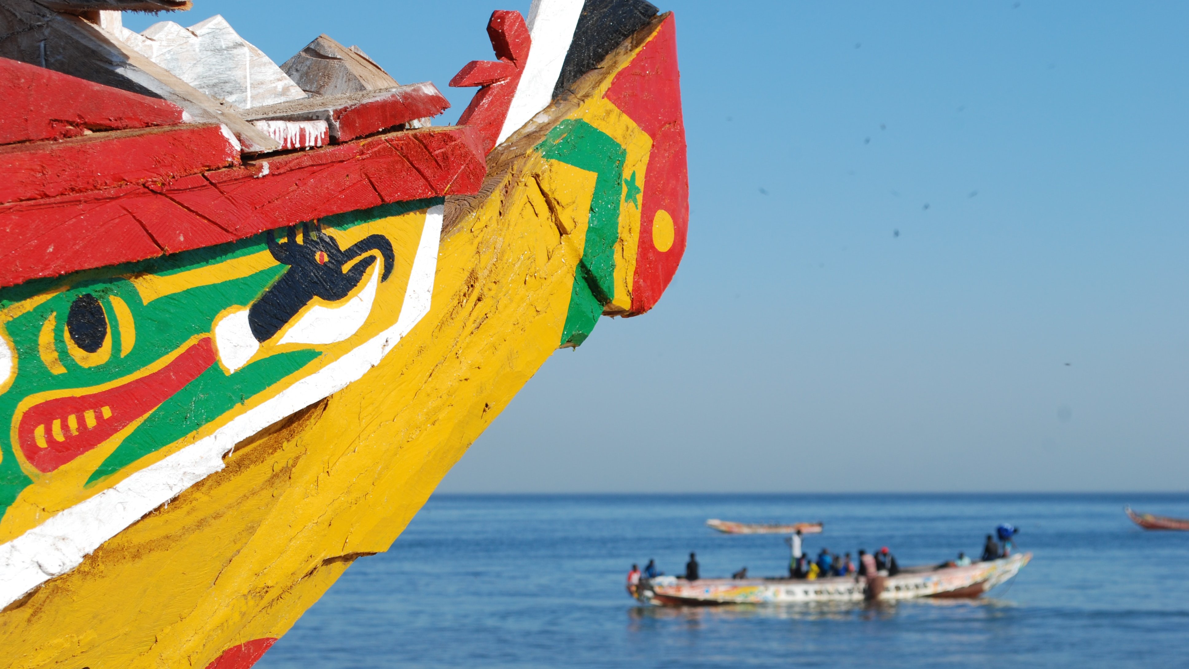 Cette image montre la proue d'une embarcation colorée, ornée de motifs vive avec principalement des couleurs rouge, jaune et verte. Les dessins évoquent des éléments symboliques, peut-être liés à la culture locale. En arrière-plan, on aperçoit d'autres bateaux sur l'eau, avec des personnes à bord, travaillant ou naviguant paisiblement. La mer apparaît calme, reflétant un ciel bleu sans nuages, créant une atmosphère sereine et ensoleillée.