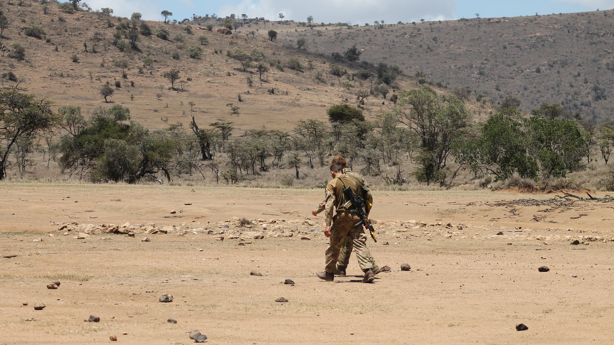 L'image montre un paysage aride et vaste, typique de la savane. Au centre, un homme marche seul sur un sol poussiéreux, parsemé de pierres. Il est vêtu d'un uniforme camouflage et porte un équipement à son dos, suggérant une activité en milieu naturel. En arrière-plan, on aperçoit des collines sèches et quelques arbres épars, témoignant d'une végétation clairsemée. Le ciel est partiellement nuageux, avec des nuages blancs qui contrastent avec le bleu du ciel. L'ensemble dégage une atmosphère de solitude et d'exploration dans un environnement sauvage.