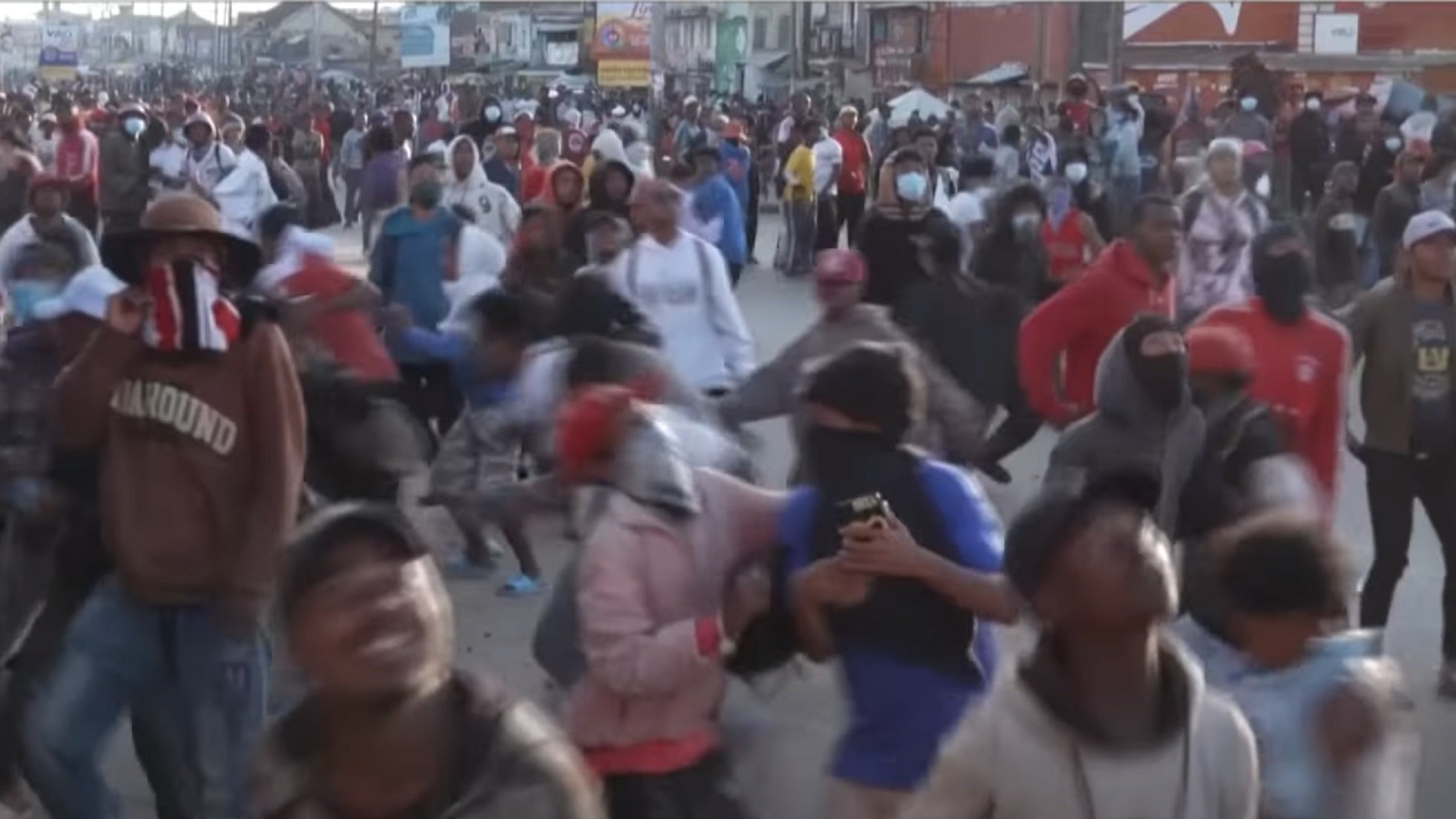 L'image montre une foule dense de personnes rassemblées dans une rue, probablement lors d'une manifestation ou d'un événement populaire. Les participants portent des vêtements variés et beaucoup d'entre eux ont des masques ou des foulards sur le visage. On peut sentir une ambiance animée et énergique, avec des mouvements rapides et des gestes expressifs. Certains individus semblent prendre des photos avec leurs téléphones. L'arrière-plan est rempli d'autres personnes, créant une atmosphère vibrante et collective.