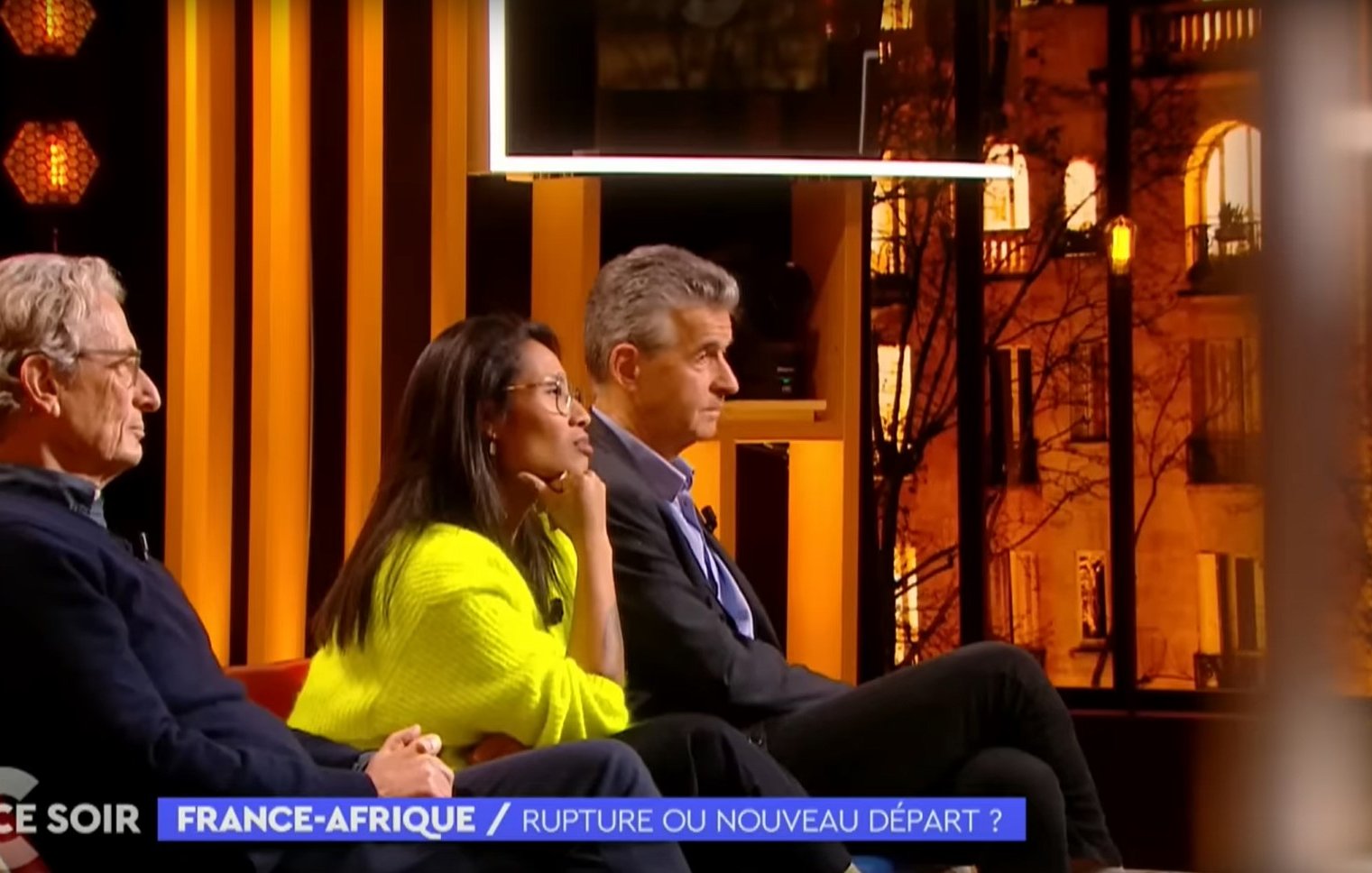 L'image montre un plateau de télévision lors d'une émission intitulée "France-Afrique : Rupture ou nouveau départ ?". Trois personnes sont assises sur un canapé, éclairées par des lumières chaudes. À gauche, un homme avec des lunettes et des cheveux grisonnants porte un pull sombre. Au centre, une femme, habillée d'un pull lumineux, semble attentive et réfléchie. À droite, un homme aux cheveux courts et argentés affiche une expression sérieuse. En arrière-plan, on peut apercevoir des fenêtres illuminées qui donnent une ambiance chaleureuse à l'ensemble. Le décor est moderne, avec des éléments graphiques en arrière-plan qui ajoutent à l'esthétique de l'émission.