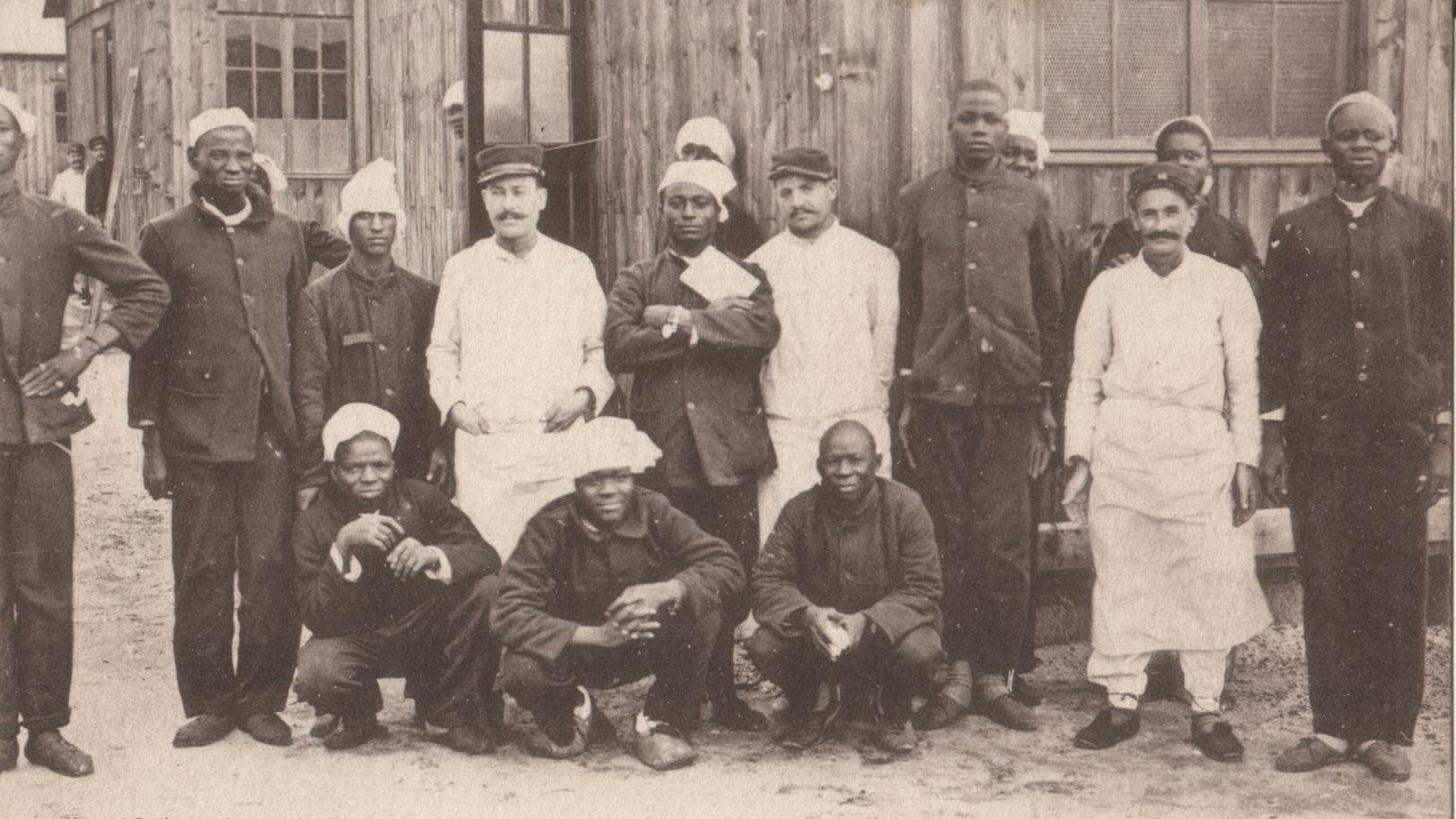 L'image montre un groupe de soldats sénégalais convalescents rassemblés devant un bâtiment en bois, vraisemblablement un hôpital de campagne. Il y a treize personnes, dont des hommes en uniforme militaire et des membres du personnel médical. Les soldats portent des uniformes sombres et certains d'entre eux ont des casquettes ou des bonnets. Le personnel médical, d'autre part, porte des blouses blanches, indiquant leur rôle. Tous affichent des expressions amicales, certains sourient tandis que d'autres prennent des poses plus sérieuses. L'arrière-plan montre des murs en bois, donnant une atmosphère de campagne ou de mobilisation militaire. Cette scène capture un moment de camaraderie et de résilience au sein des soldats pendant une période difficile.