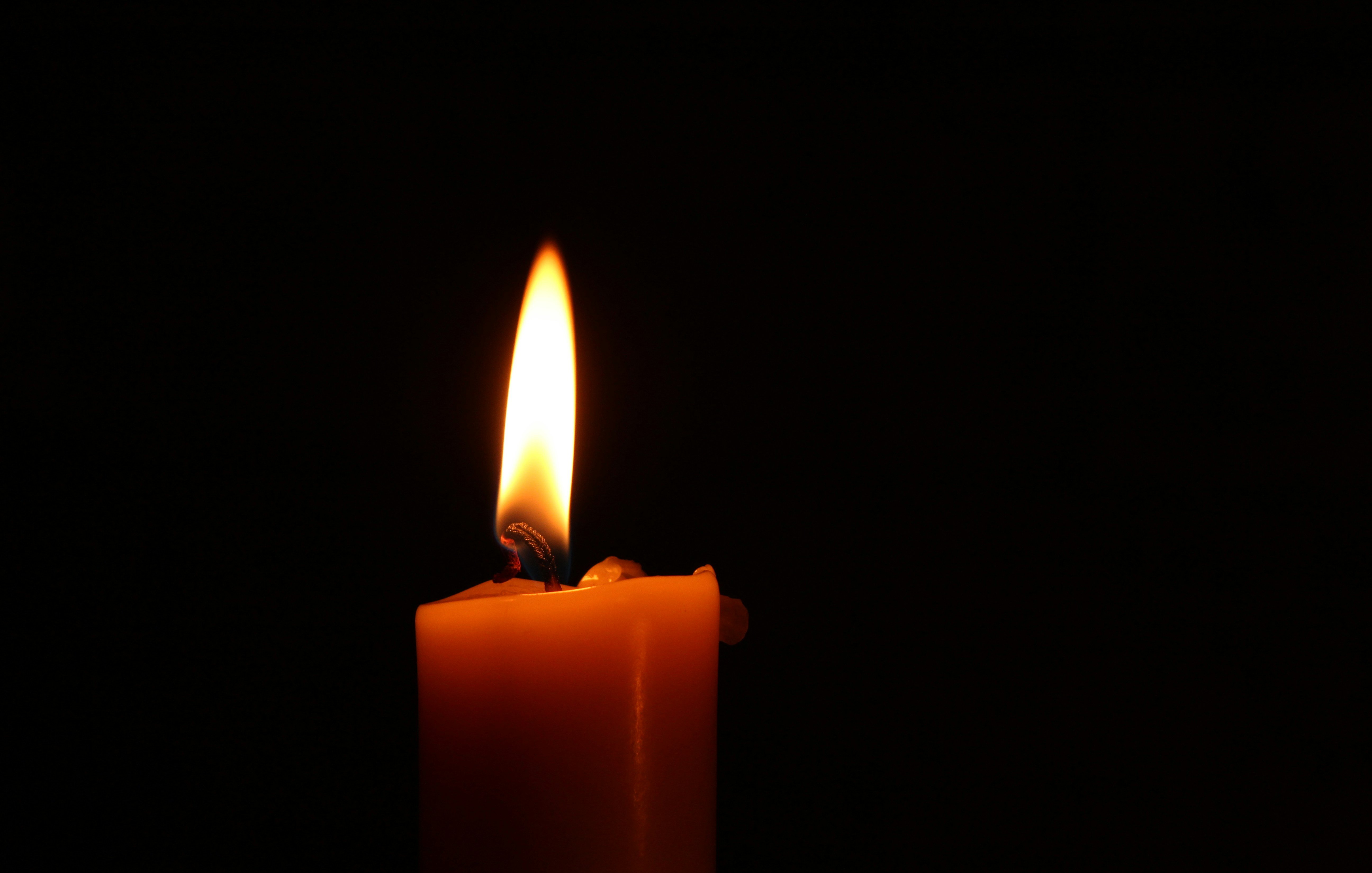 L'image présente une bougie allumée dans un environnement sombre. La flamme, vive et dansante, émet une lumière chaleureuse et douce, créant une atmosphère calme et apaisante. La surface de la bougie est lisse et de couleur jaune ou orange, et la mèche, légèrement recourbée, est en train de se consumer doucement. La lumière de la flamme projette des ombres délicates autour, accentuant l'obscurité environnante. Cette scène évoque une sensation de sérénité et de chaleur, typique d'un moment de méditation ou de recueillement.