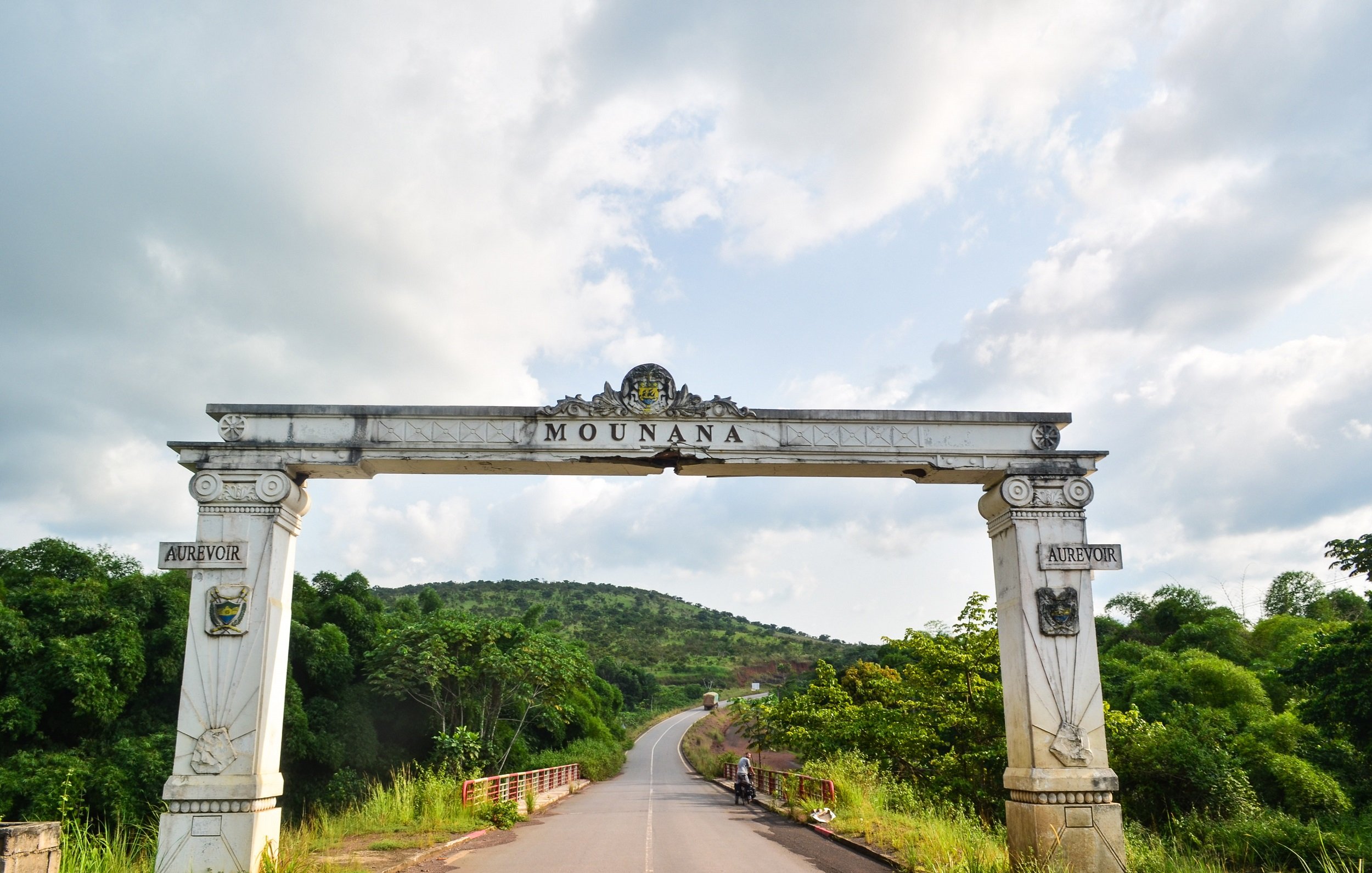 L'image présente une grande arche située à l'entrée de la ville de Mounana. Cette arche, ornée de motifs architecturaux, est peinte en blanc avec des détails dorés et armoriaux de chaque côté. Au-dessus de l'entrée, le nom "MOUNANA" est inscrit en lettres stylisées. En arrière-plan, on aperçoit un paysage verdoyant avec des collines et une route qui serpente entre des arbres. Le ciel est partiellement nuageux, créant une atmosphère paisible et accueillante. L'ensemble évoque l'entrée d'un lieu significatif, invitant les visiteurs à découvrir ce qui se trouve au-delà de l'arche.