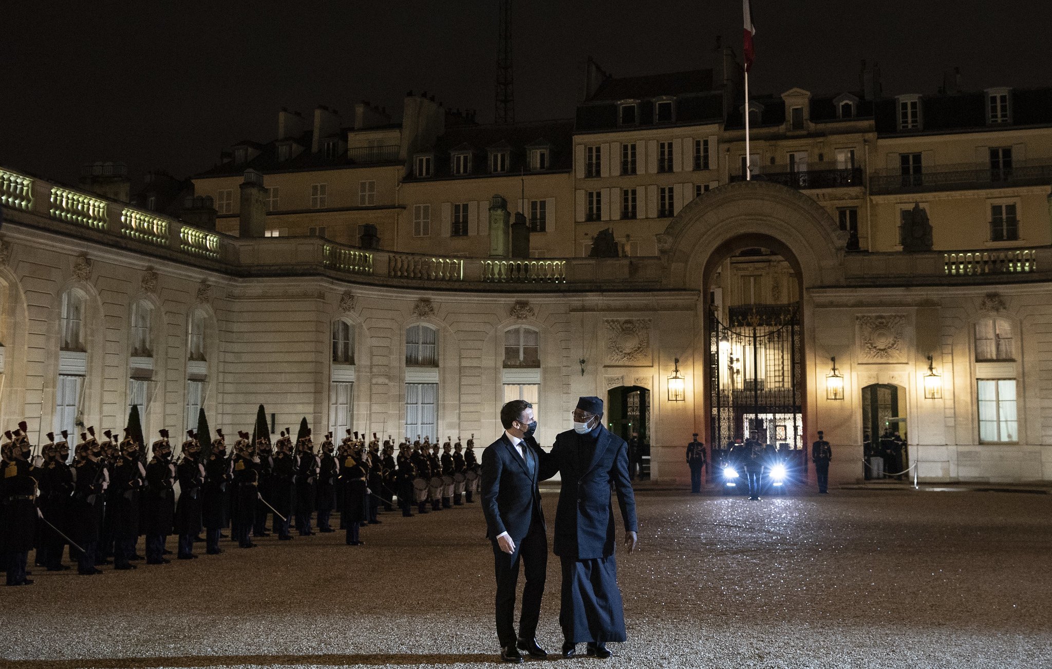 L'image représente une scène nocturne au Palais de l'Élysée, en France. Au premier plan, deux hommes discutent, l'un portant un costume noir et l'autre vêtu d'un long manteau sombre. En arrière-plan, une rangée de soldats en uniforme se tient en position, formant une haie d'honneur. Les bâtiments classiquement architecturés du palais sont illuminés, mettant en valeur leurs détails. Une lumière provenant d'un véhicule est visible à droite, ajoutant à l'atmosphère solennelle du moment. Les drapeaux français flottent au-dessus du bâtiment. L'ensemble de la scène dégage une impression de formalité et de dignité.