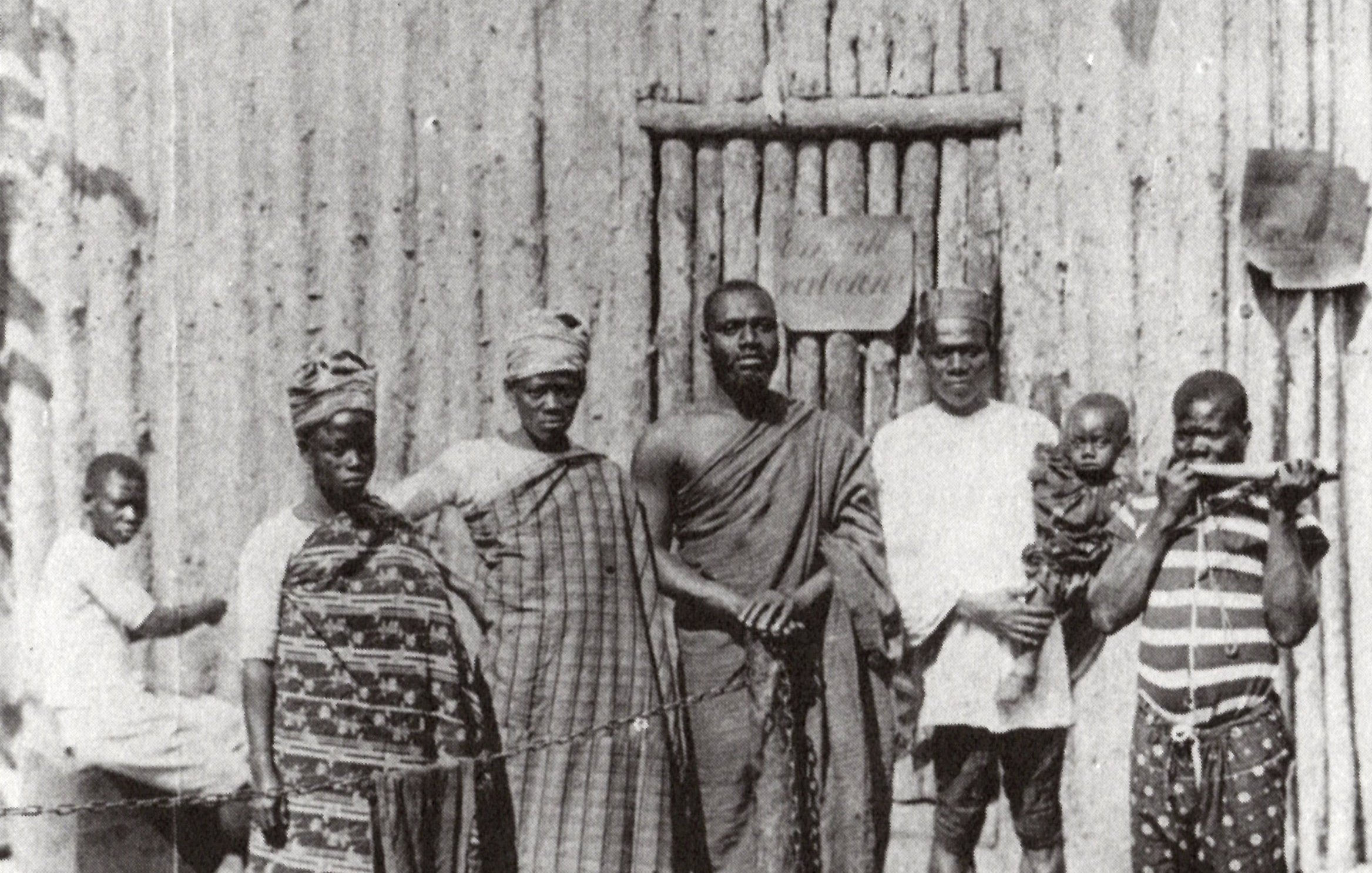 L'image montre un groupe de six personnes se tenant debout devant une structure en bois, qui semble être une maison ou un abri, avec un décor rustique. Au fond, on distingue un homme assis qui fait face à la scène principale. Les membres du groupe portent des vêtements traditionnels typiques, avec des motifs variés, affichant une richesse culturelle. L'un d'eux tient un enfant, tandis qu'un autre est équipé d'un bâton, suggérant une certaine autorité ou dignité. L'atmosphère de l'image dégage un sentiment de communauté et de convivialité.