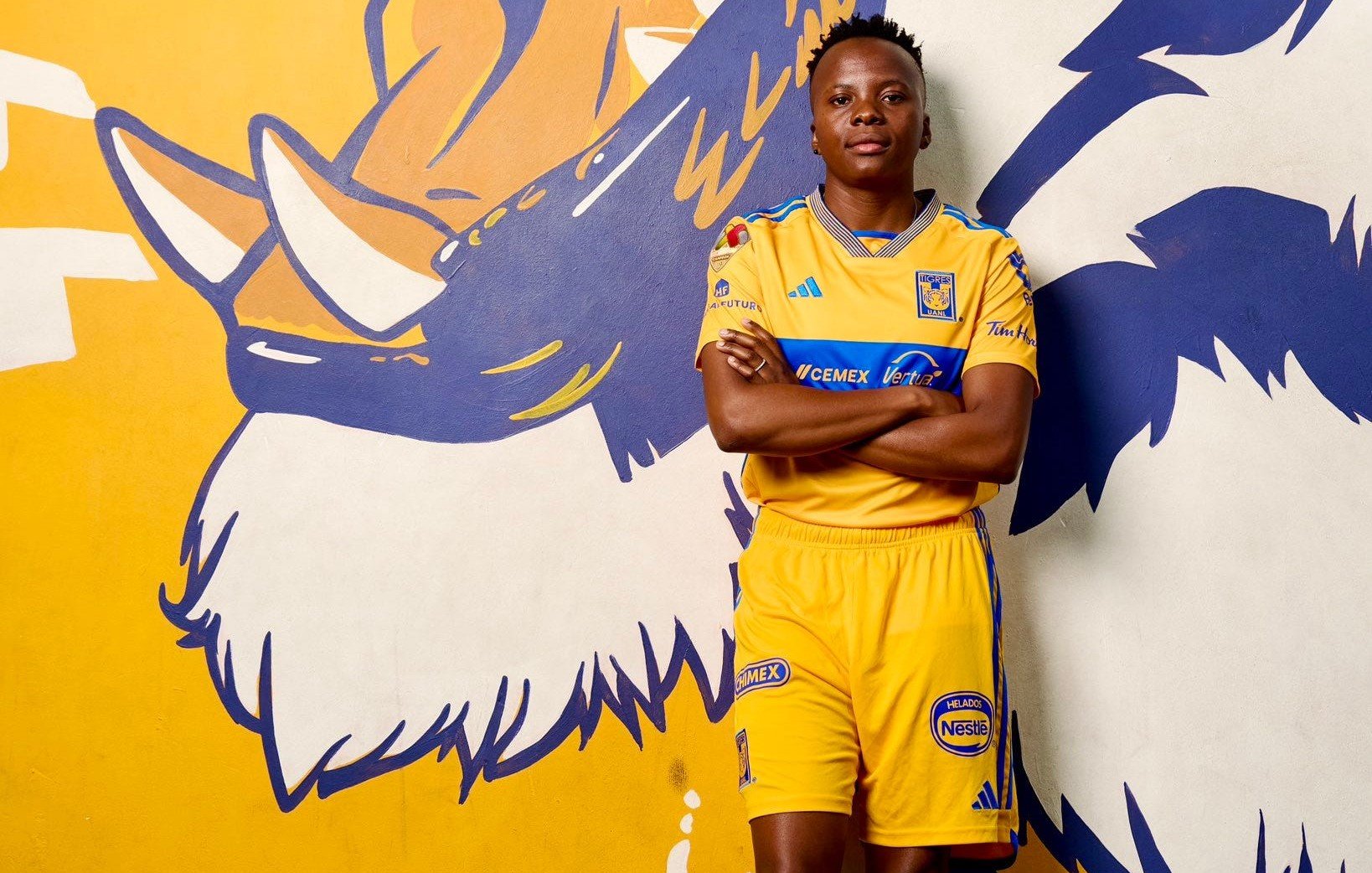 L'image montre un jeune homme debout devant un mur peint en jaune et bleu, qui représente un tigre rugissant. Il porte un uniforme de footbal aux couleurs vives, principalement jaune, avec des détails bleus. Son attitude est confiante ; il se tient les bras croisés, affichant une expression de détermination. Le mur qui l'entoure ajoute une dynamique visuelle avec des motifs de griffes et des éléments stylisés qui renforcent le thème du tigre. Le sol est recouvert d'herbe artificielle, évoquant un terrain de sport.