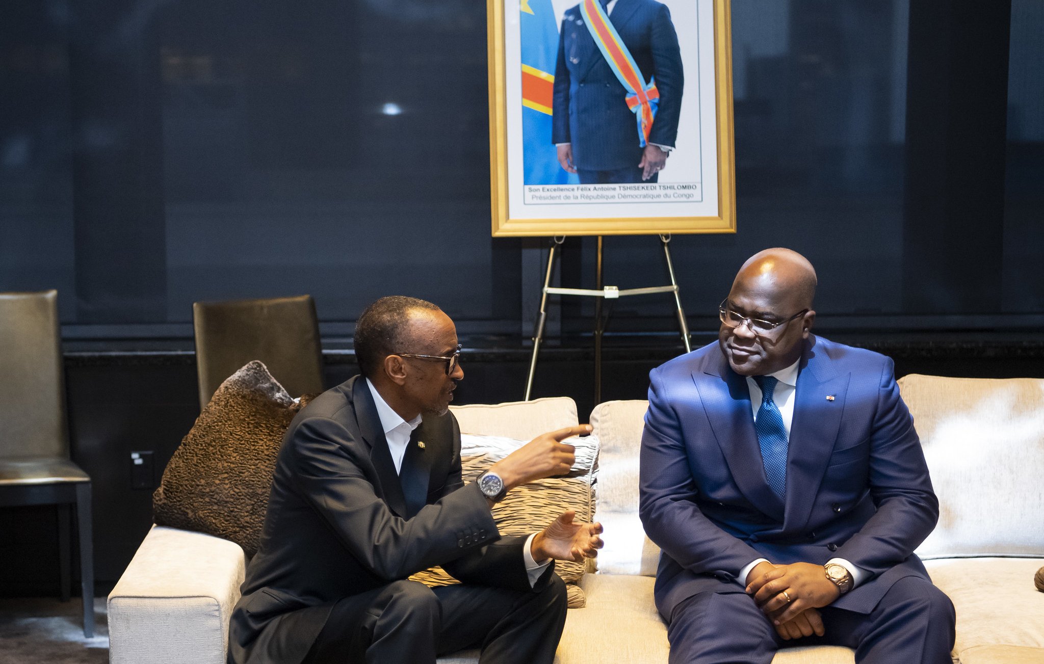 L'image montre deux hommes assis sur un canapé dans un cadre intérieur élégant. L'un des hommes porte un costume noir et semble s'engager dans une conversation. L'autre homme est habillé d'un costume bleu et sourit. En arrière-plan, il y a un portrait encadré d'un homme en costume, avec un drapeau de la République démocratique du Congo visible derrière lui. L'environnement est moderne, avec des luminaires doux et un décor soigné, suggérant une ambiance formelle ou diplomatique.
