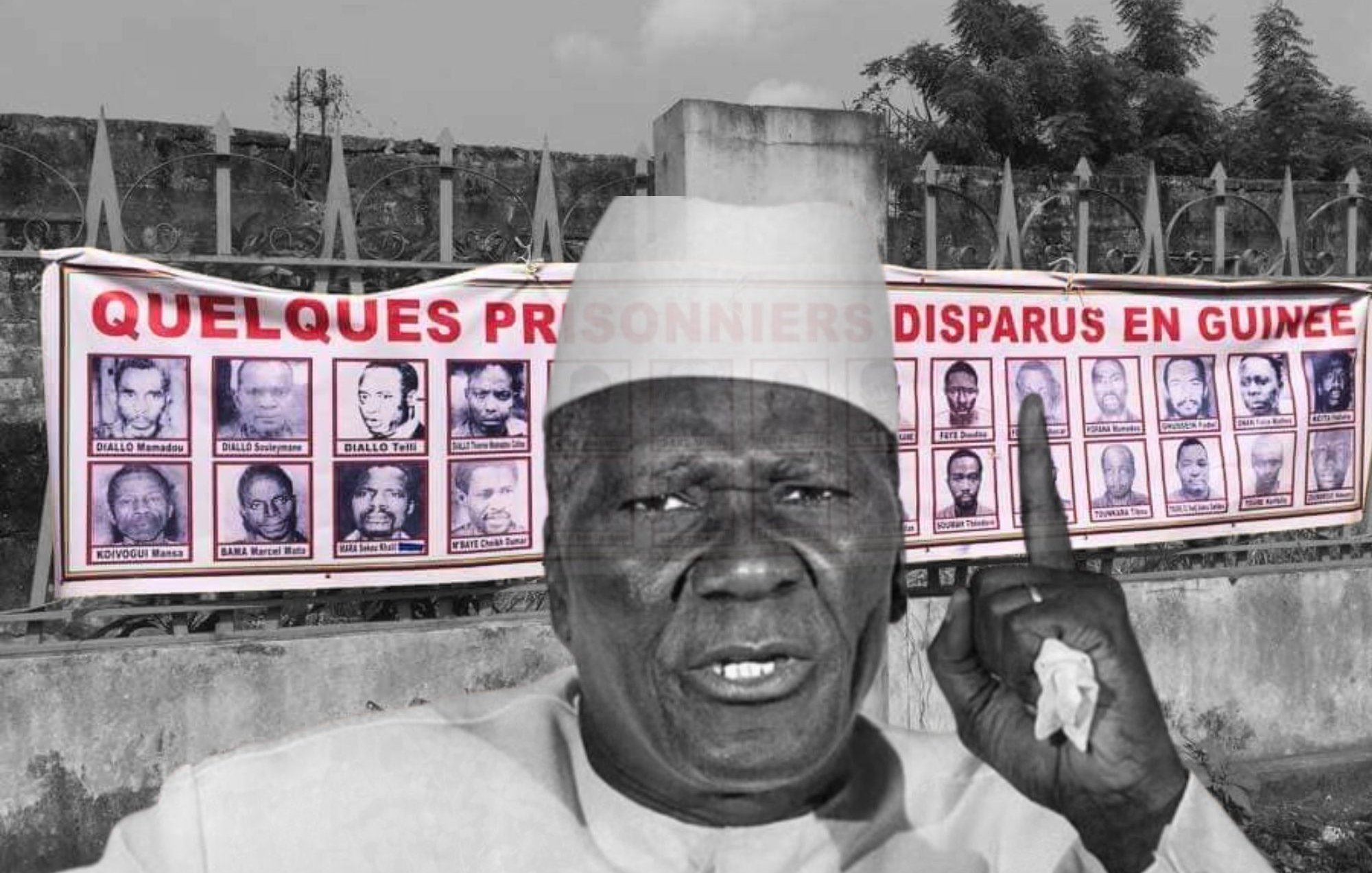 L'image montre un homme d'âge mûr, vêtu d'une tenue traditionnelle et portant un chapeau. Il lève un doigt en signe d'alerte ou de demande d'attention. En arrière-plan, on peut voir une grande banderole accrochée sur un mur. Cette banderole affiche des portraits de plusieurs personnes, accompagnés d'un texte qui fait référence à des prisonniers disparus en Guinée. L'ambiance de l'image évoque un appel à ne pas oublier ces disparitions et à sensibiliser le public à cette problématique.