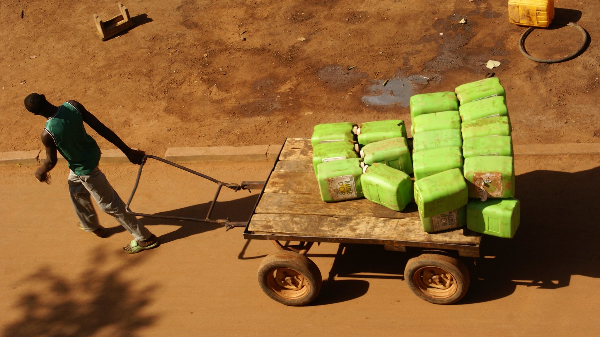 L'image montre un homme poussant une charrette en bois sur une route en terre. La charrette est remplie de nombreuses bouteilles vertes, probablement des contenants d'eau. L'homme porte un T-shirt sans manches et un pantalon gris, et son corps semble penché en avant, indiquant l'effort physique qu'il fournit. Le sol autour de lui est sec et légèrement poussiéreux, avec quelques traces visibles montrant que d'autres véhicules ou personnes ont déjà passé. En arrière-plan, il pourrait y avoir des objets comme des seaux ou des tuyaux, mais l'accent est mis sur l'homme et sa cargaison. La lumière est forte, suggérant une journée ensoleillée.