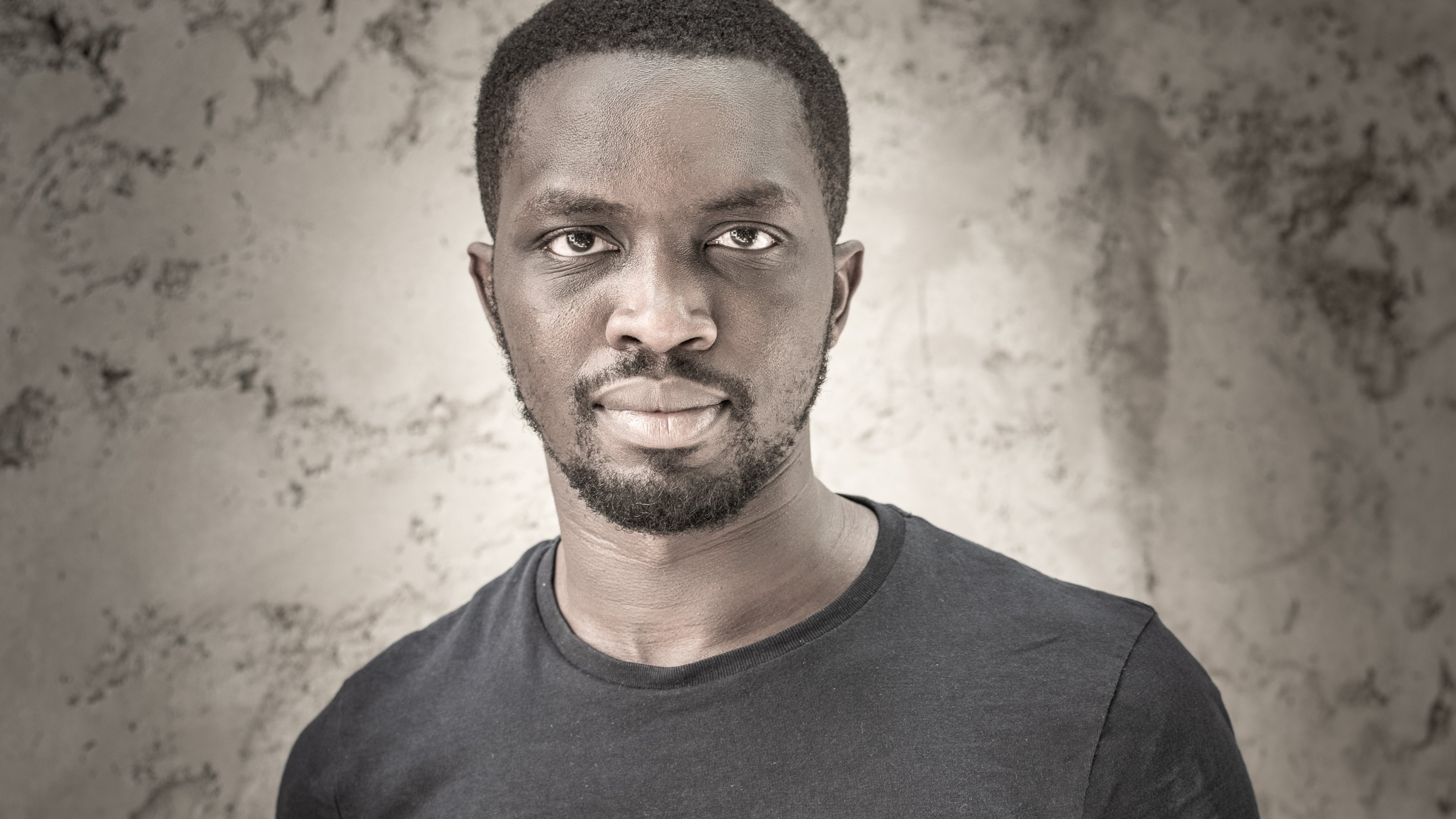 Cette image présente un homme debout, portant un t-shirt noir. Il a la peau foncée et un style de coiffure court. Son expression est sérieuse, et il regarde directement vers l'objectif avec une intensité qui attire l'attention. Le fond de l'image est texturé et neutre, mettant en valeur son visage et son corps. La lumière éclaircit certaines zones, ajoutant de la profondeur et du contraste à l'ensemble.