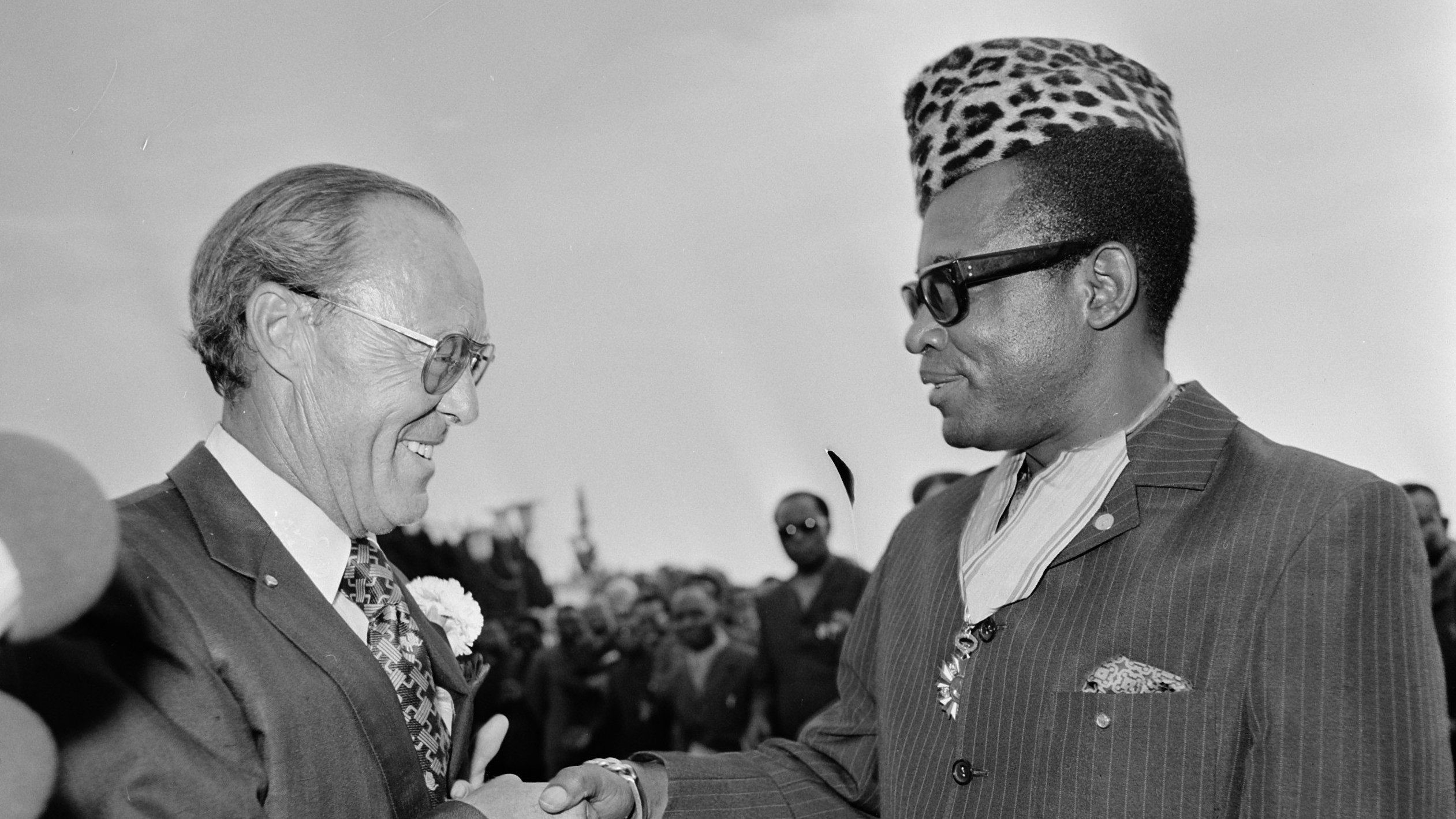 L'image montre une scène de rencontre entre deux hommes souriants, se serrant la main dans un cadre formel et convivial. Le premier homme porte un costume sombre avec une chemise à motifs fleuris et des lunettes, tandis que le second, qui semble être une figure importante, arbore un élégant costume rayé et un chapeau à motifs distinctifs, évoquant une riche culture. En arrière-plan, un groupe de personnes observe la scène, ajoutant à l'impression d'un événement important. L'atmosphère est chaleureuse et amicale, suggérant une rencontre diplomatique ou un événement marquant.