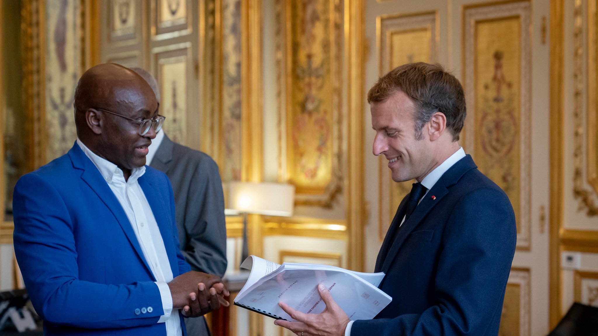 L'image montre deux hommes se tenant debout dans une pièce élégante et ornée. L'un des hommes porte un costume bleu vif et sourit tandis qu'il parle. L'autre homme, vêtu d'un costume sombre, tient des documents dans ses mains et lui répond avec un léger sourire. La décoration de la pièce est luxueuse, avec des murs dorés et des éléments classiques qui évoquent un cadre formel et historique. Il y a une lumière chaleureuse, créant une ambiance conviviale à cet échange.