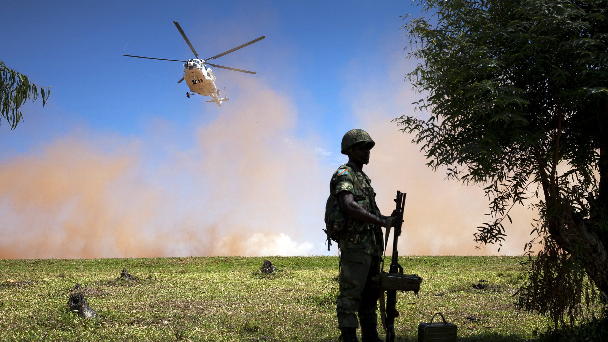 L'image représente un paysage militaire. Au premier plan, on voit un soldat debout, tenant une arme. Il porte un casque et un uniforme camouflage, suggérant qu'il se trouve dans un environnement opérationnel. À l'arrière-plan, un hélicoptère survole la scène, produisant un mouvement et une dynamique. Le ciel est clair, mais il y a également un nuage de poussière ou de fumée, ajoutant une atmosphère dramatique à la scène. Le terrain est verdoyant, mais des éléments comme des troncs d'arbres ou des obstacles peuvent être perçus, contribuant à l'ambiance militaire et à l'action en cours.