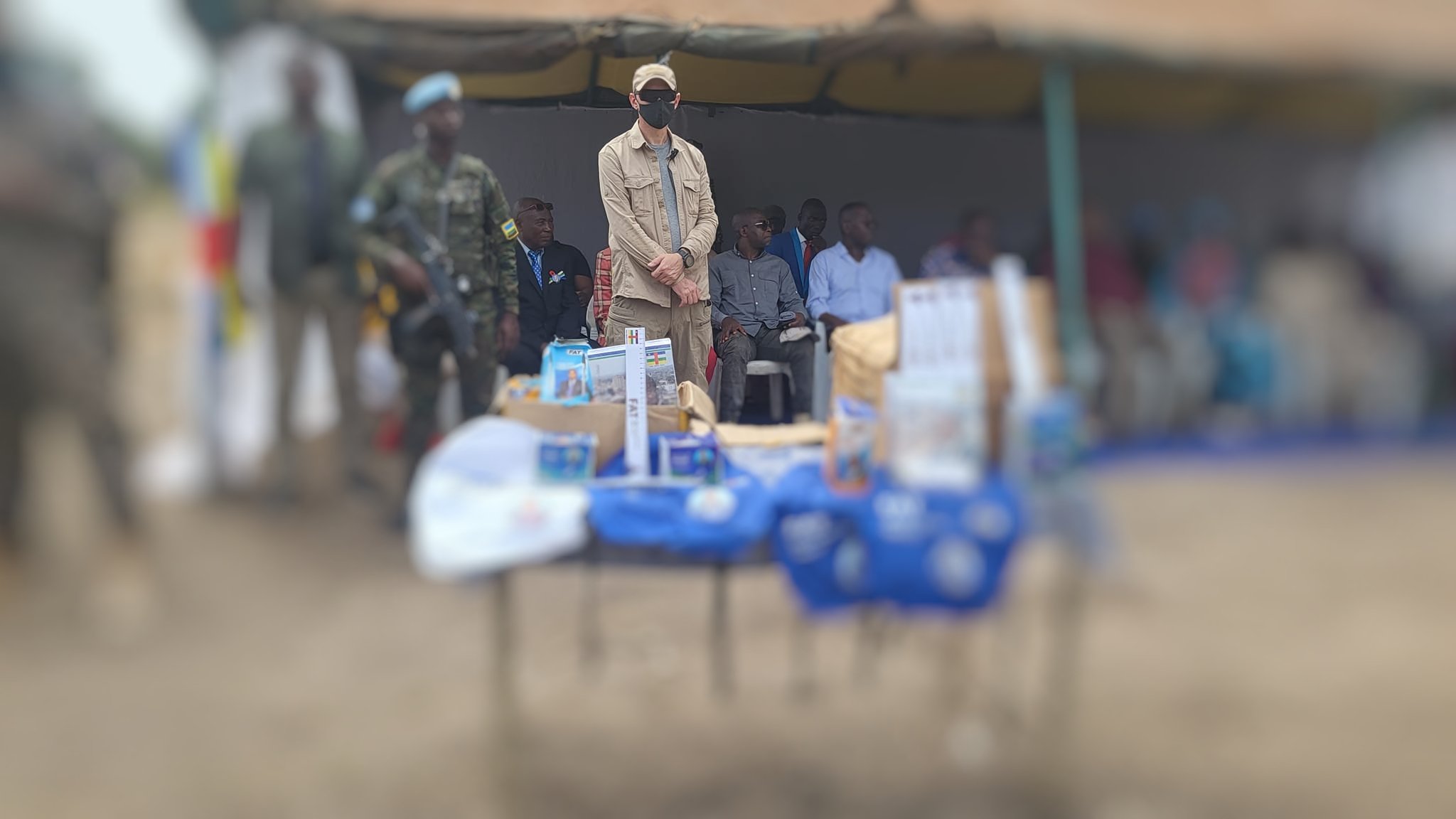 L'image montre un événement en plein air, avec une tente sur le côté. Au centre, on voit une table couverte d'objets et de brochures, probablement liés à une présentation ou un programme. À l'arrière-plan, plusieurs personnes sont assises, y compris des enfants et des adultes. Un homme se tient debout, portant une casquette et un masque, semblant être un intervenant ou une personne d'autorité. La scène reflète un moment de rassemblement communautaire, chargé d'une atmosphère d'échange et d'information.