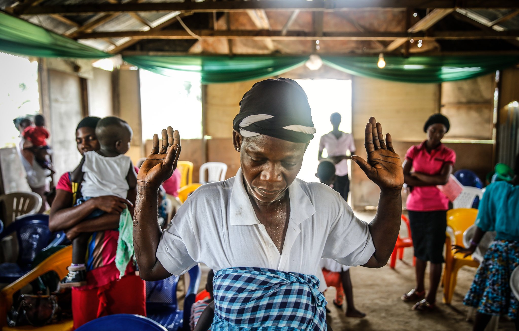 L'image montre un intérieur de salle, où des chaises colorées sont disposées de manière informelle. Au premier plan, une femme se tient debout, les bras levés, dans une posture de prière ou de méditation. Elle porte un foulard sur la tête et une blouse blanche, tandis qu'une jupe à motifs colorés entoure sa taille. En arrière-plan, on aperçoit d'autres personnes, certaines sont engagées dans une conversation tandis que d'autres semblent également en prière. L'ambiance est empreinte de spiritualité et d'un sentiment de communauté. La lumière naturelle entre par des fenêtres, créant un espace à la fois chaleureux et intime.