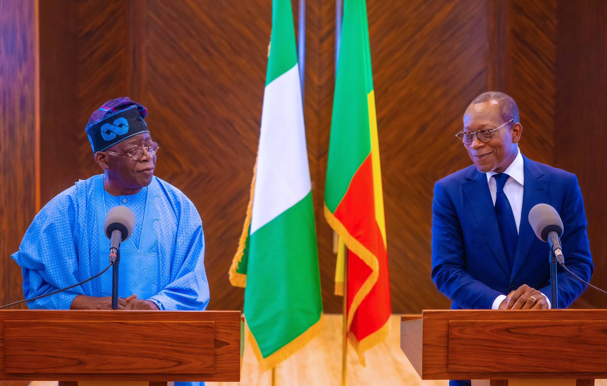 L'image montre deux hommes se tenant devant des pupitres en bois dans un cadre officiel. À leur arrière-plan, on peut voir les drapeaux du Nigéria (vert, blanc, vert) et du Bénin (rouge, jaune, vert). L'homme à gauche porte une tenue traditionnelle de couleur bleue avec un chapeau, tandis que l'homme à droite est habillé en costume sombre. Les deux hommes semblent engagés dans une conversation, exprimant une atmosphère de dialogue et de coopération. Le décor est élégant, soulignant le caractère officiel de la rencontre.