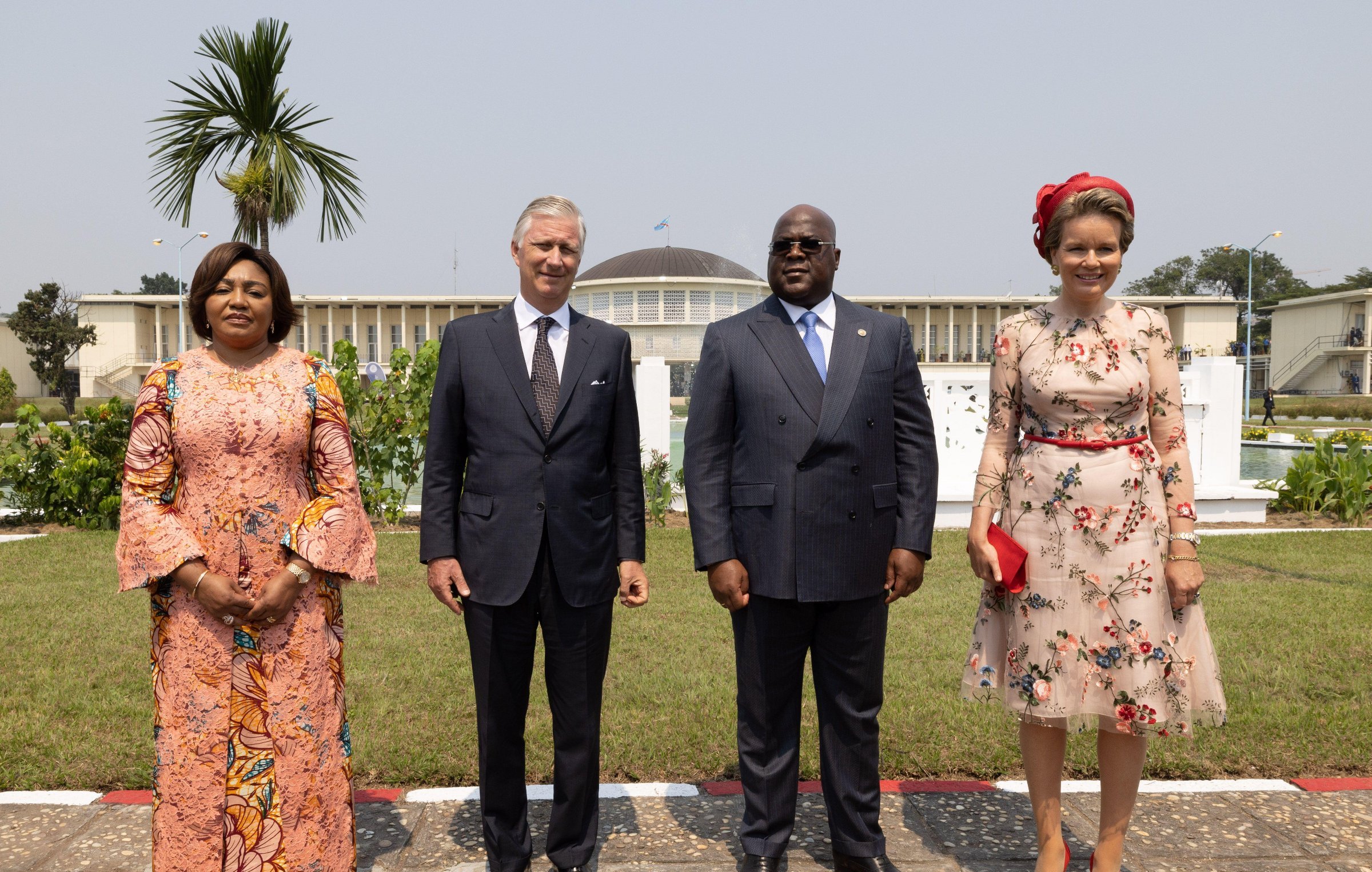 L'image montre un groupe de quatre personnes se tenant debout dans un jardin bien entretenu. À gauche, une femme porte une robe longue à motifs floraux et des manches évasées. À côté d'elle, un homme en costume sombre est debout, affichant une expression chaleureuse. Ensuite, un autre homme, également en costume, a une posture amicale et est au centre de l'image. Finalement, à droite, une femme porte une robe à fleurs avec un joli chapeau assorti et un sac à main rouge, se tenant avec élégance. En arrière-plan, on aperçoit un bâtiment imposant et un palmier qui ajoutent une atmosphère exotique à la scène. Le ciel est clair, ce qui contribue à un sentiment de rencontre officielle.