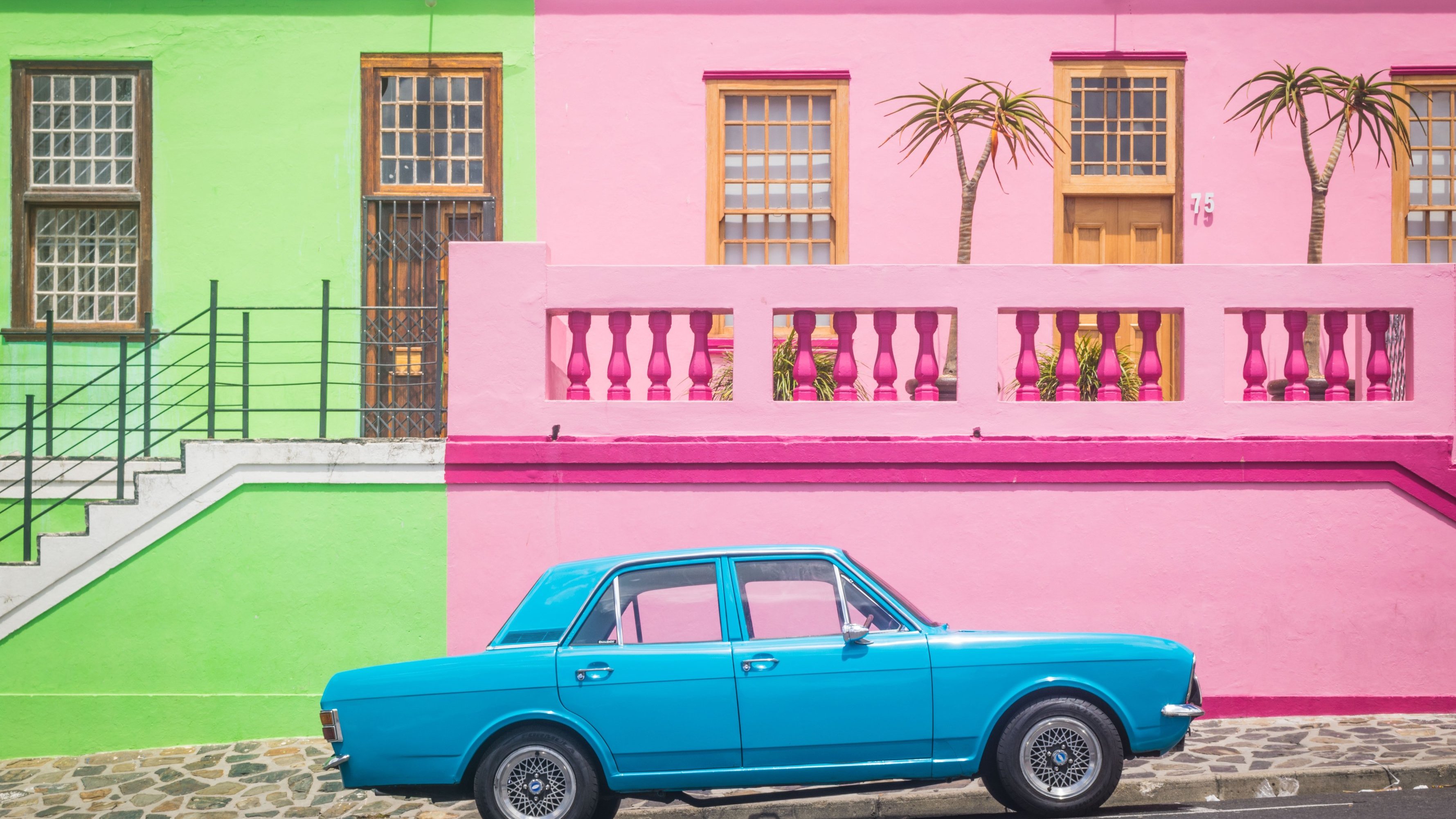 L'image présente une scène vibrante et colorée d'une rue typique. À gauche, un bâtiment se dresse en vert clair, avec des fenêtres en bois et un accès par escalier. À droite, un mur rose vif forme la façade d'une maison, ornée de deux palmiers qui ajoutent une touche tropicale. Entre les deux bâtiments, une voiture de couleur bleu clair est garée, se détachant joliment sur le fond coloré. Le sol est pavé, et l'ambiance générale de la scène est joyeuse et accueillante, évoquant une atmosphère estivale et ensoleillée.