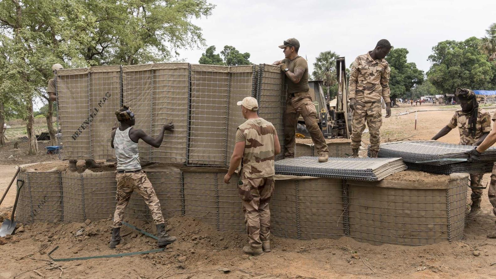 L'image montre un groupe de soldats au travail sur un site de construction. Ils sont en train de manipuler de grandes structures en treillis, qui semblent être remplies de terre. L'environnement est naturel, avec des arbres verts en arrière-plan et un sol légèrement argileux. Les soldats portent des uniformes militaires et semblent concentrés sur leur tâche. Certains d'entre eux soulèvent des éléments, tandis que d'autres les positionnent soigneusement. Cette scène évoque une atmosphère de collaboration et d'effort collectif.