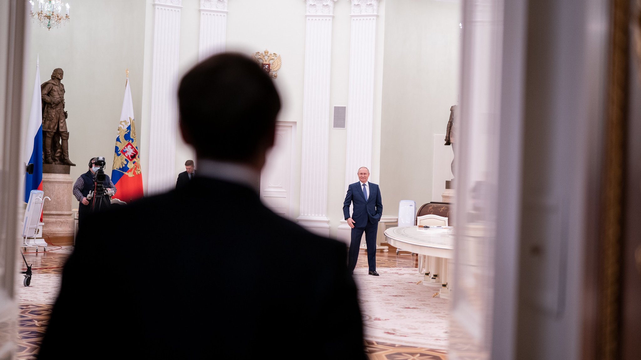 L'image montre une scène dans un intérieur officiel, probablement un palais ou un bureau gouvernemental. Au premier plan, on voit le dos d'une personne en costume sombre, qui semble faire face à une autre personne située à une certaine distance. Cette deuxième personne, qui est clairement identifiable comme une figure politique importante, se tient debout dans une pièce au décor élégant, avec une lumière douce. À l'arrière-plan, on peut distinguer des drapeaux russe et peut-être d'autres éléments symboliques. Le sol est en marbre, et les murs sont peints dans des teintes claires, ajoutant une ambiance solennelle à l'ensemble. L'atmosphère de l'image suggère un moment de rencontre ou de dialogue important.