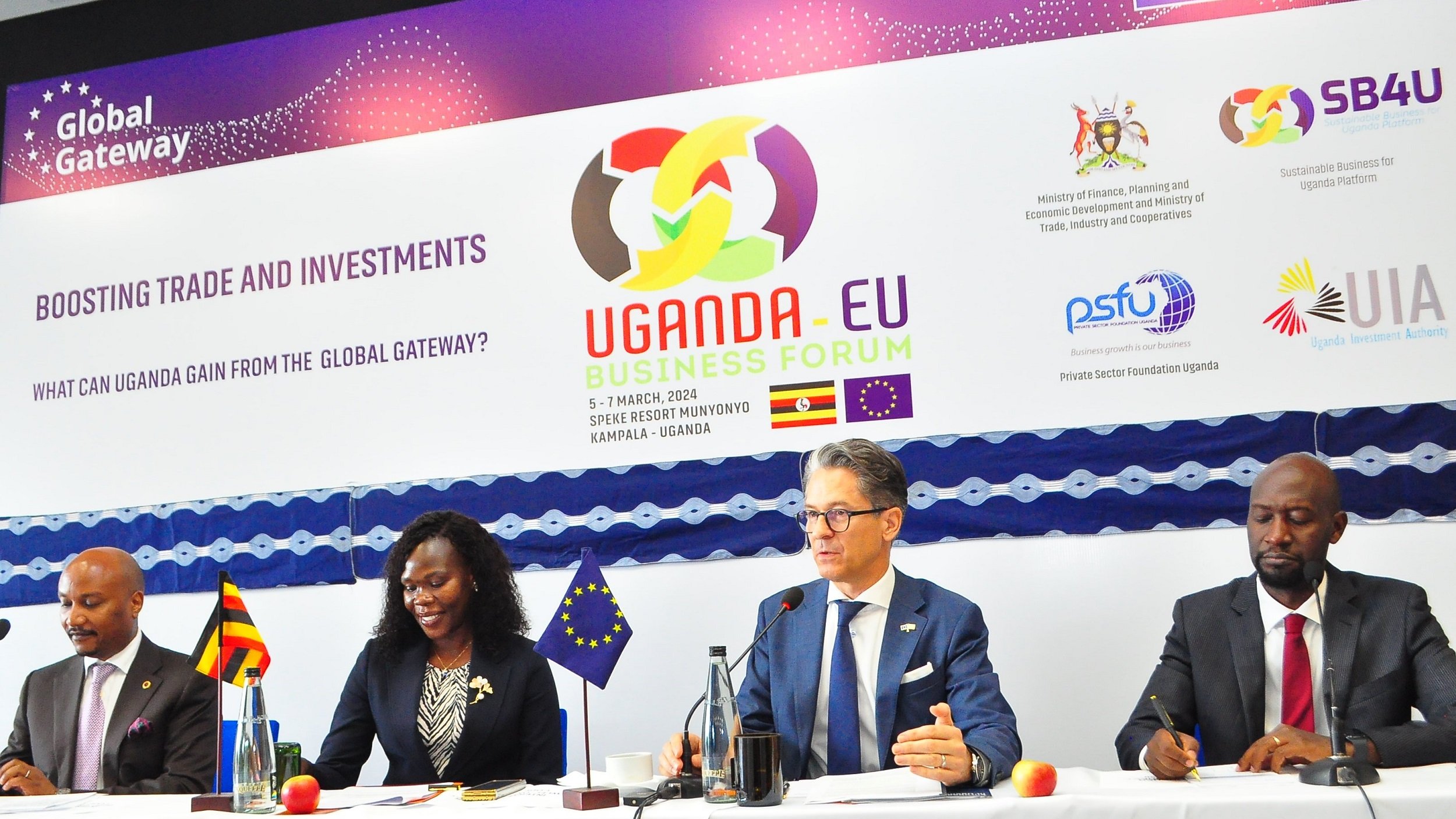 L'image montre une scène d'un forum d'affaires intitulé "Uganda-EU Business Forum". Au premier plan, quatre personnes sont assises à une table, chacune ayant un micro devant elle. À gauche, un homme souriant semble représenter l'Ouganda. Ensuite, une femme, également souriante, prend la parole. Au centre, un homme, avec des lunettes et un air sérieux, est en train de s'exprimer. Enfin, à droite, un autre homme, avec une posture attentive, écoute. En arrière-plan, une banderole avec des logos et le titre de l'événement est accrochée. L'ambiance est professionnelle, et l'événement semble axé sur le renforcement des échanges et des investissements entre l'Uganda et l'Union européenne.