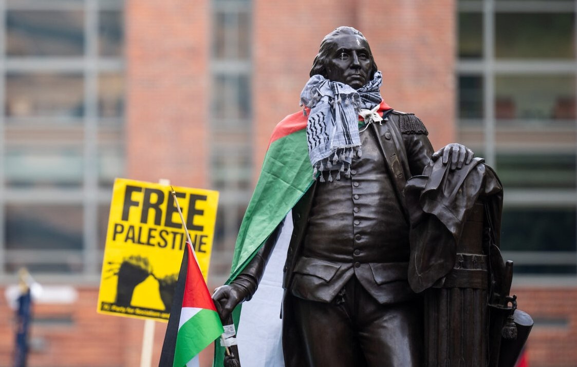 L'image montre une statue en bronze d'un homme debout, tenant un bâton dans une main et un document dans l'autre. Cette statue est habillée d'un keffieh, un foulard traditionnel palestinien en noir et blanc, et elle est drapée d'un drapeau palestinien au motif rouge, blanc, noir et vert. En arrière-plan, on aperçoit un panneau jaune sur lequel est inscrit "FREE PALESTINE" en lettres noires, exprimant un message de soutien pour la cause palestinienne. L'environnement semble être une place publique animée, avec des bâtiments en briques visibles derrière. Cette scène témoigne de la mobilisation et de la solidarité, créant une ambiance engageante et militante.