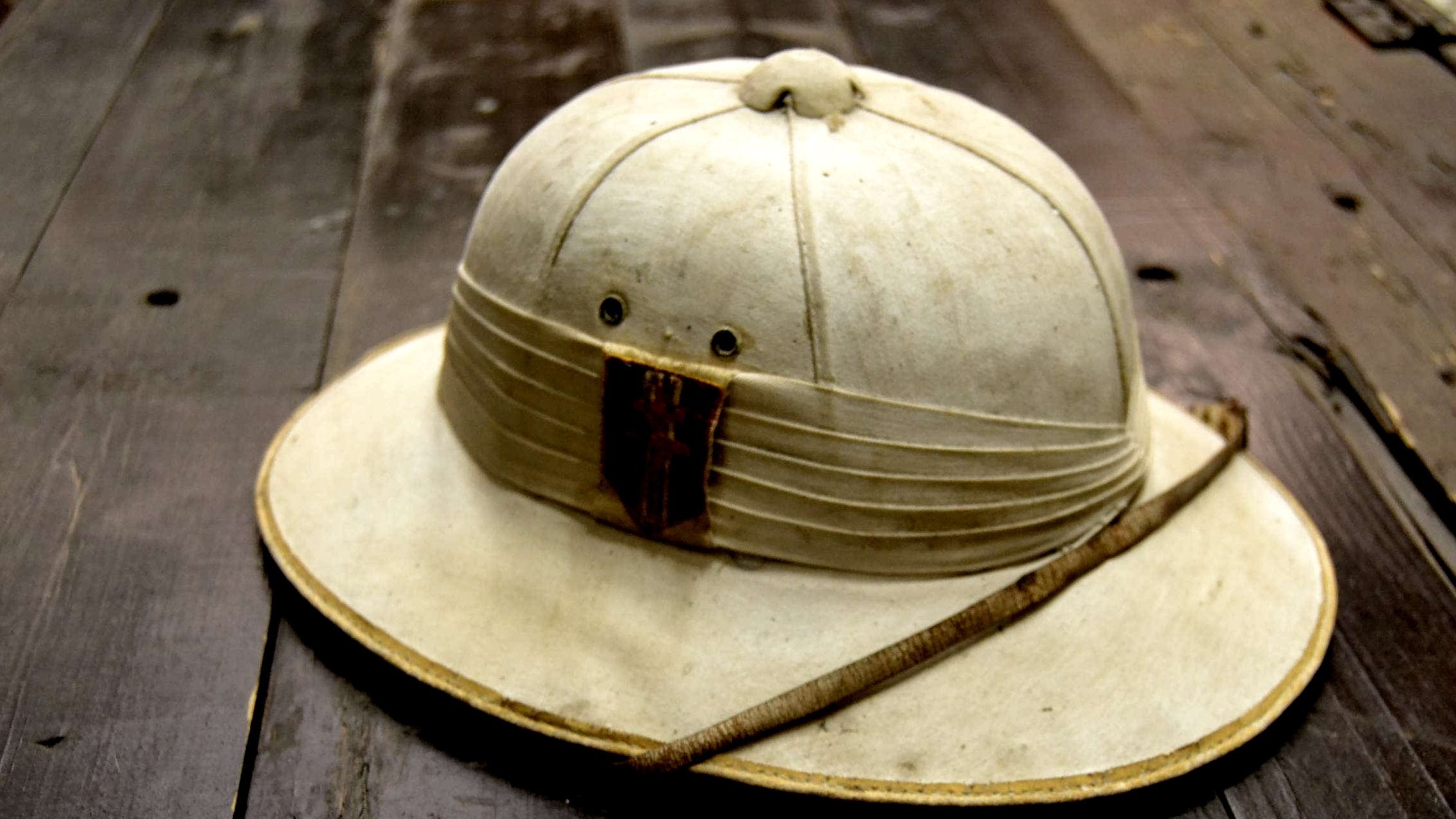 L'image montre un chapeau de style explorer, souvent associé aux expéditions dans des régions chaudes ou tropicales. Le chapeau est de couleur claire, presque beige, avec une large bordure qui protège du soleil. Sa partie supérieure est légèrement surélevée, indiquant une forme arrondie. À l'avant, il y a une bande de tissu plissé avec un morceau de bois ou de cuir, donnant un aspect rustique. Une fine tige est posée sur le chapeau, ajoutant une touche naturelle. La surface du chapeau semble légèrement usée, témoignant d'un usage antérieur. Le fond sur lequel il repose est en bois, ce qui renforce l'atmosphère d'aventure.